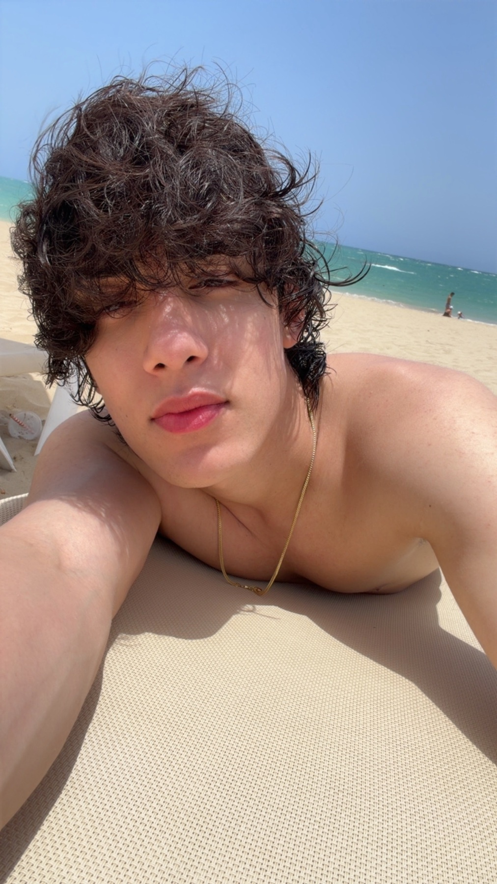 A young man with curly dark hair and tan skin takes a selfie on a beach chair. He is wearing a gold chain and appears to be enjoying a sunny day by the ocean.