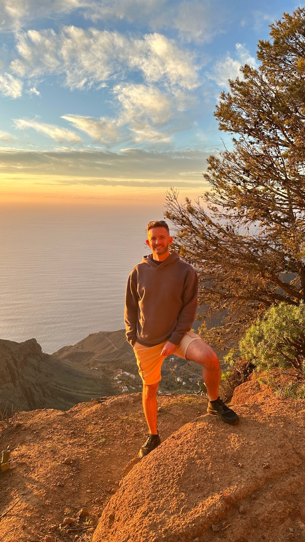A man stands on a rocky cliff overlooking the ocean at sunset. He is wearing a grey hoodie and tan shorts, with a warm, golden light illuminating the scene.
