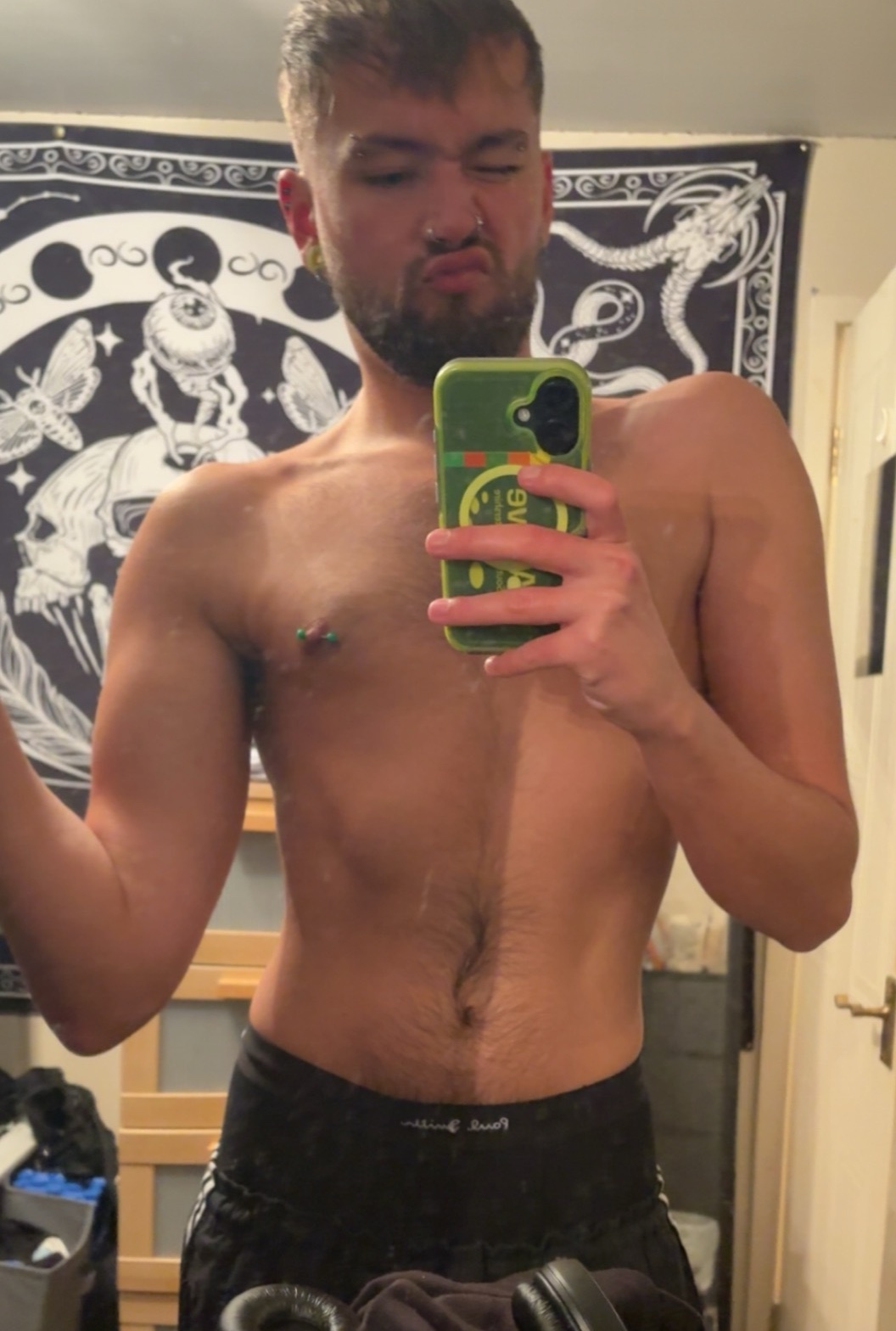 A shirtless man takes a mirror selfie. He has dark hair, stubble, and a chest piercing.