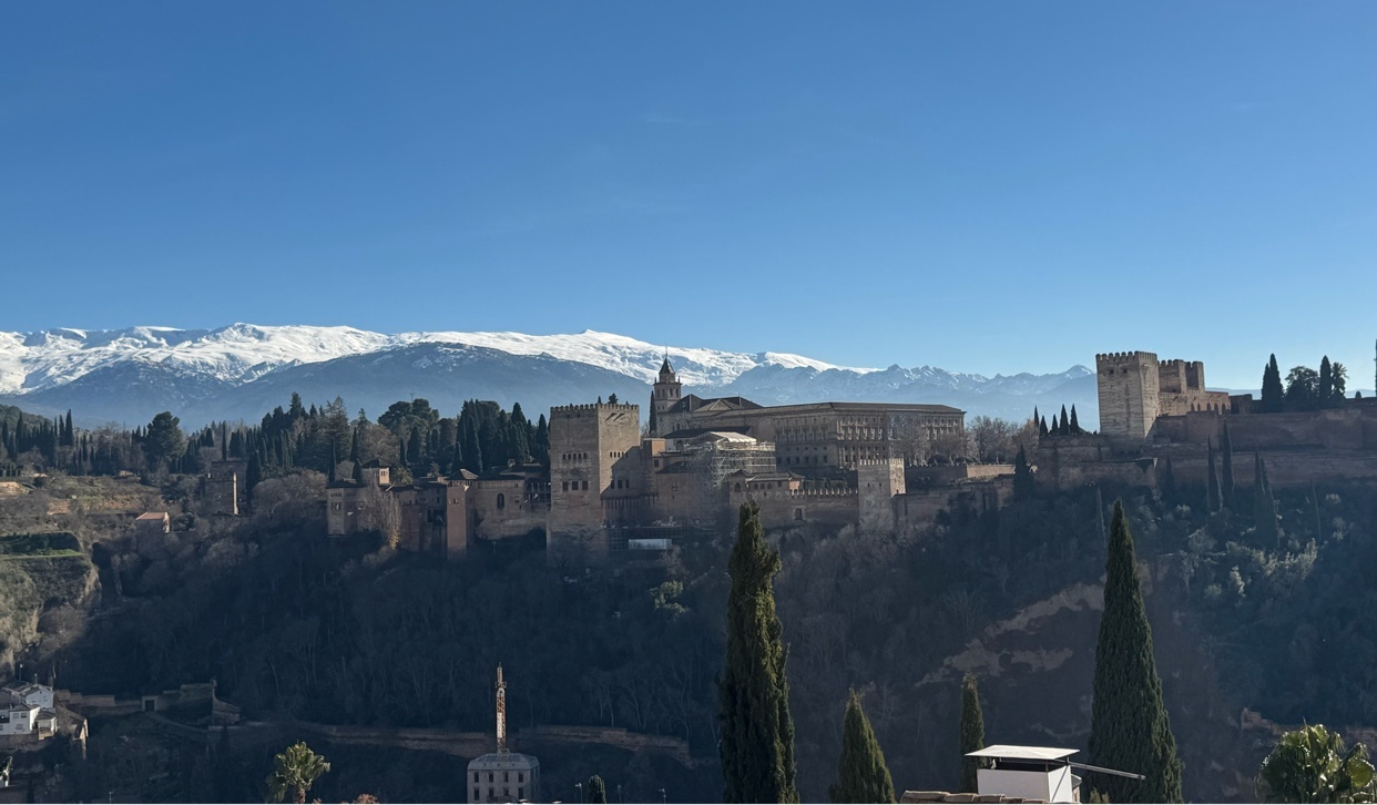 A scenic view of the Alhambra in Granada, Spain, with snow-capped mountains in the background. The image captures the historical architecture against a clear blue sky.