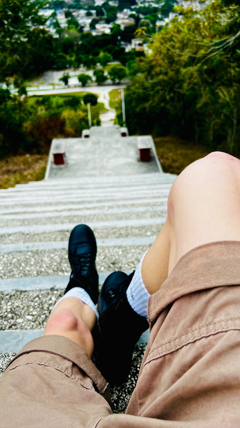 A person's legs are shown from the knees down, sitting on a set of outdoor stairs. The person is wearing shorts, socks, and black sneakers, with a view of a park and distant buildings behind them.