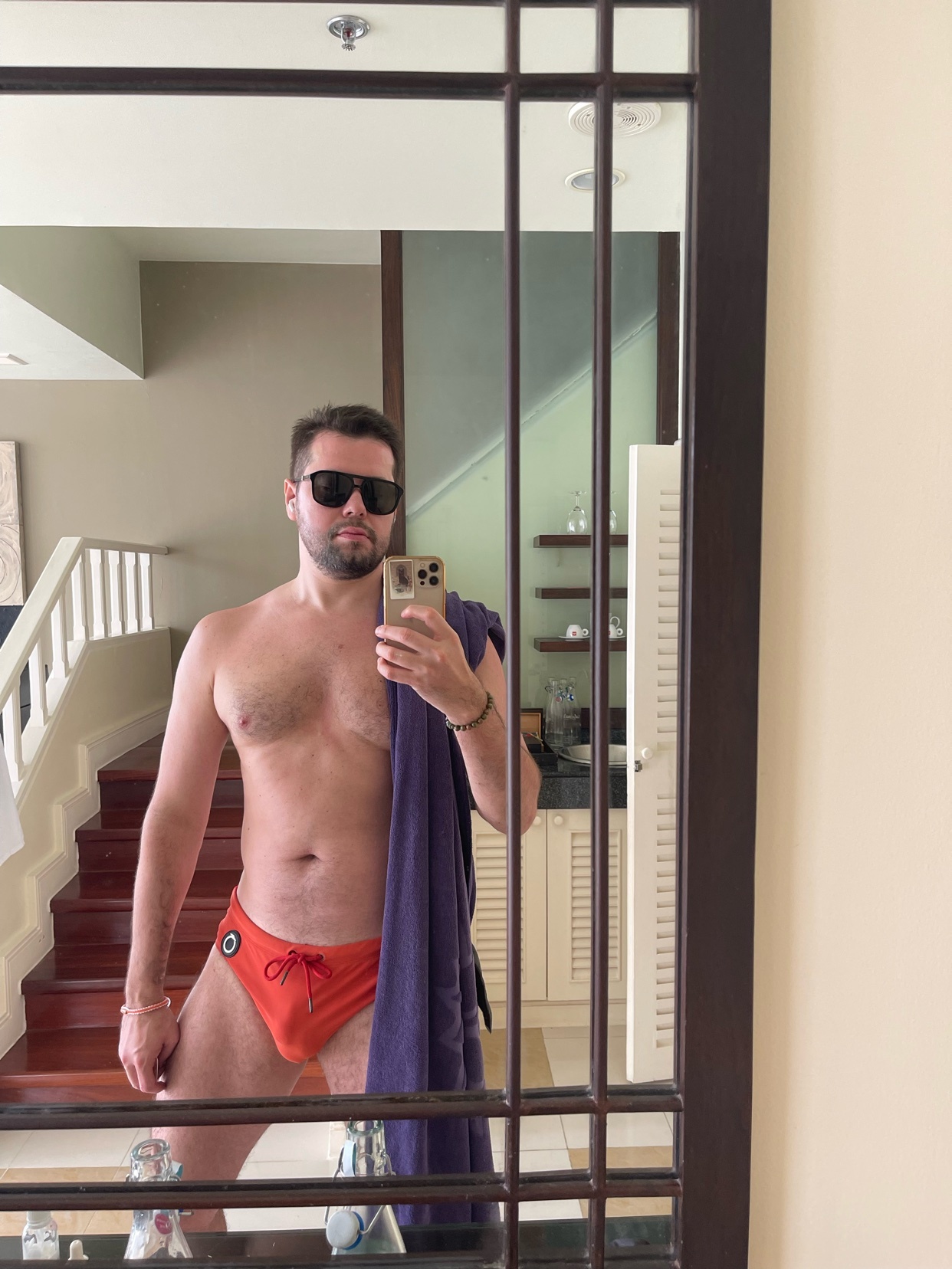 A man takes a mirror selfie, shirtless and wearing orange swimwear. He is holding a phone and a towel.
