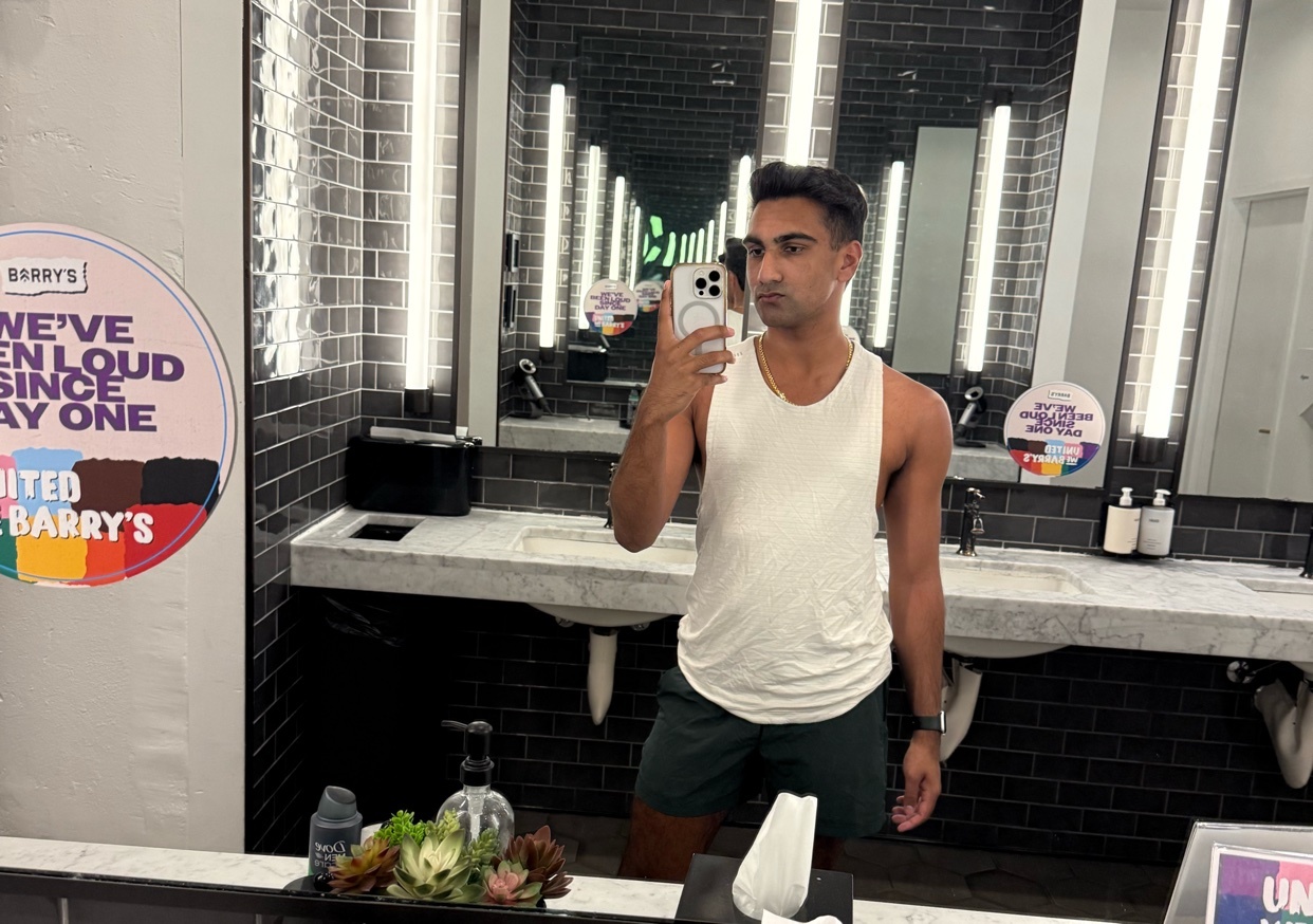 A man takes a mirror selfie in a gym bathroom. He is wearing a white tank top and dark shorts, and the background shows a row of sinks and mirrors.