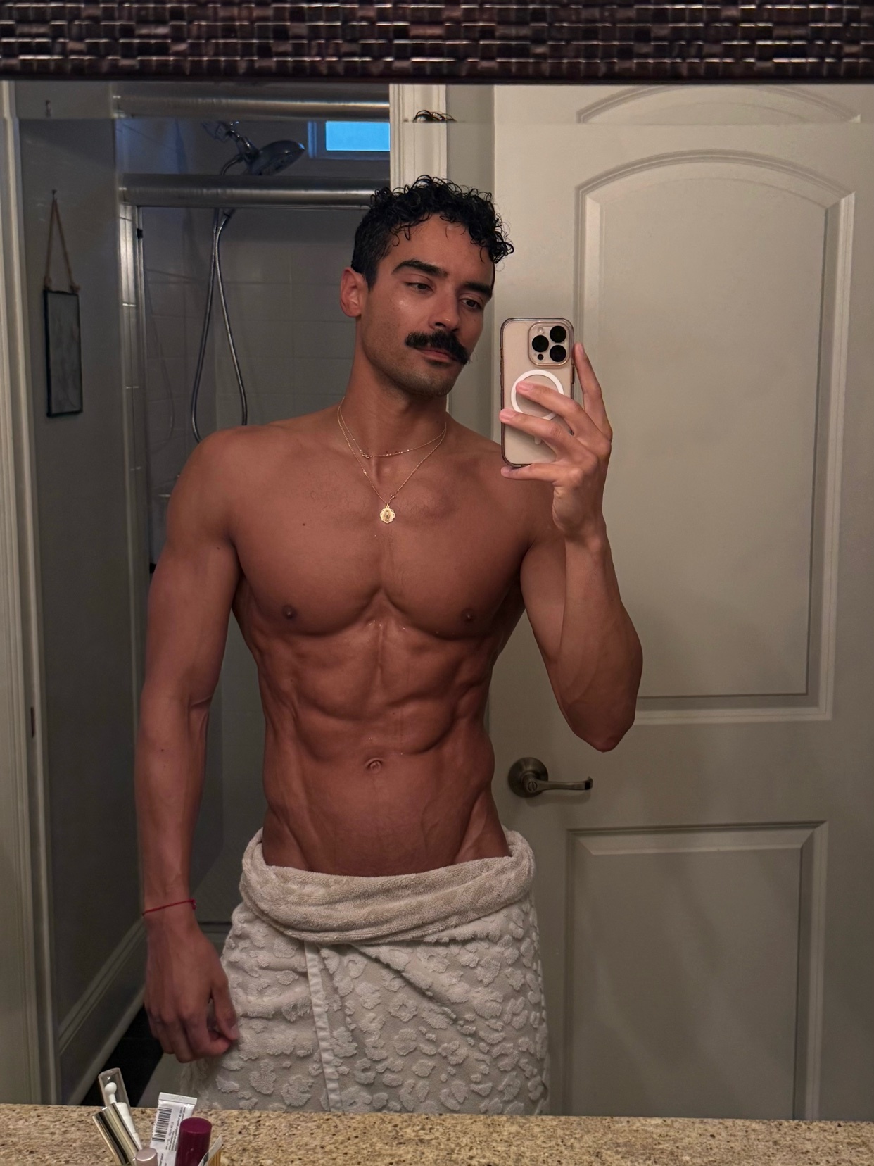 A man with a mustache takes a mirror selfie in a bathroom, wearing a towel around his waist and showing off his abs.