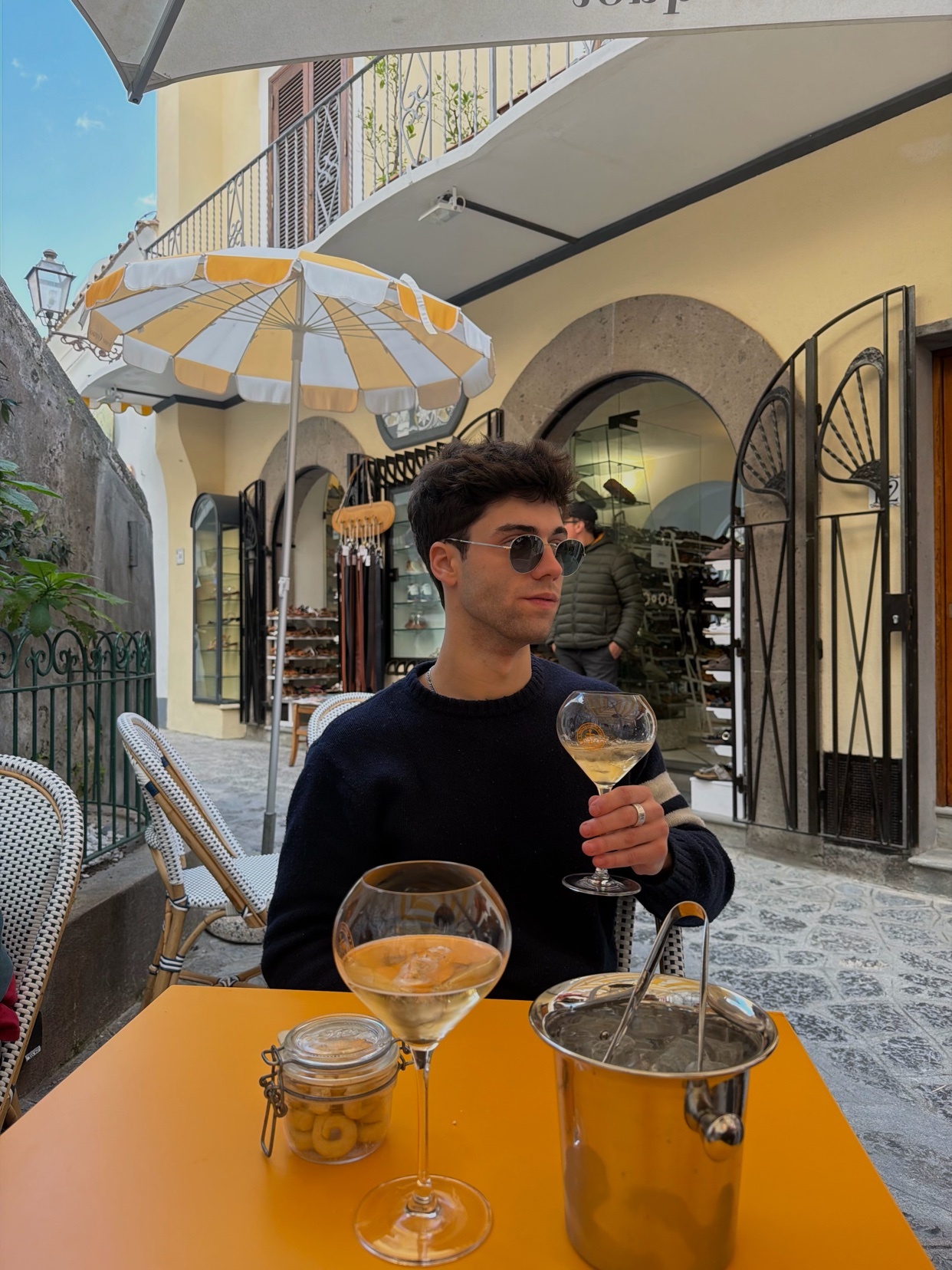 A man sits at an outdoor cafe table with a glass of white wine and an ice bucket. He wears sunglasses and a dark sweater, looking off to the side.