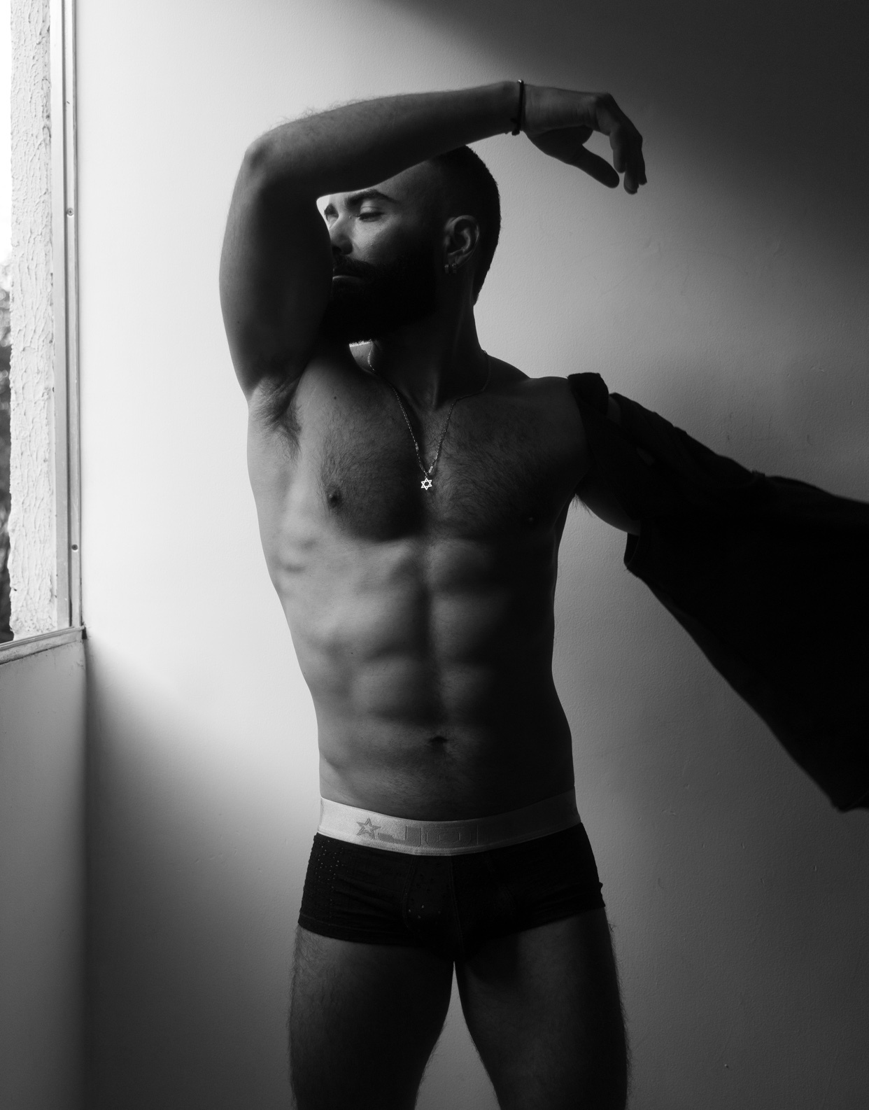 A black and white photo of a man in underwear, showing off his physique. He is standing near a window, with his arm raised.