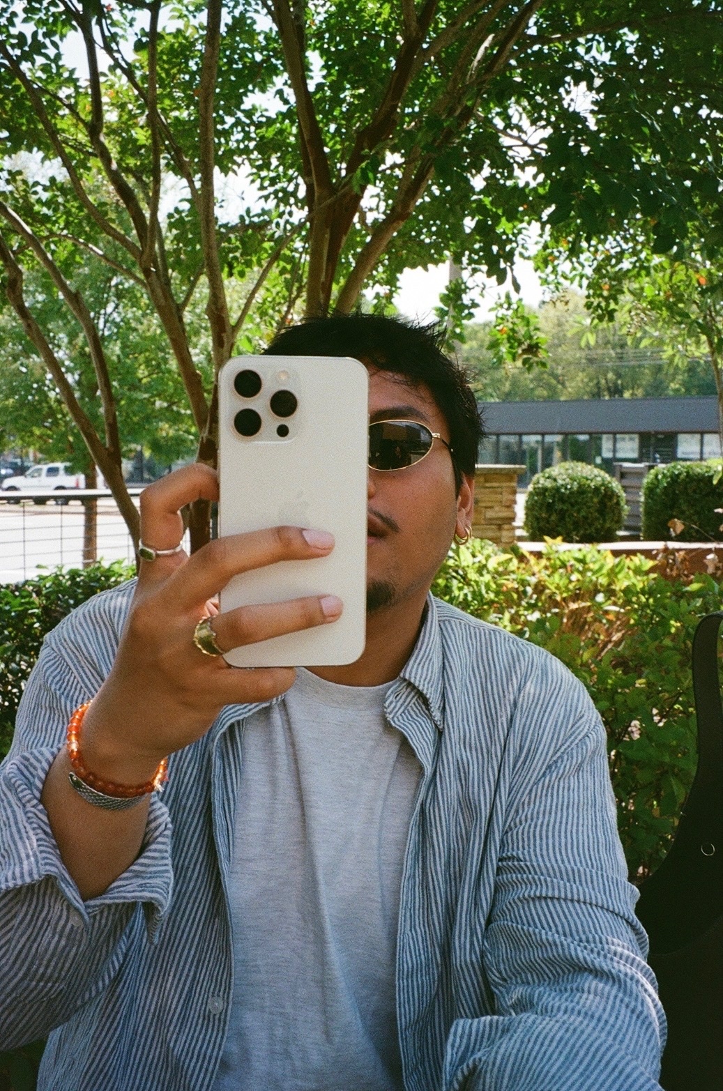 A man takes a selfie with his phone, partially obscuring his face. He wears sunglasses, a button-down shirt, and rings.