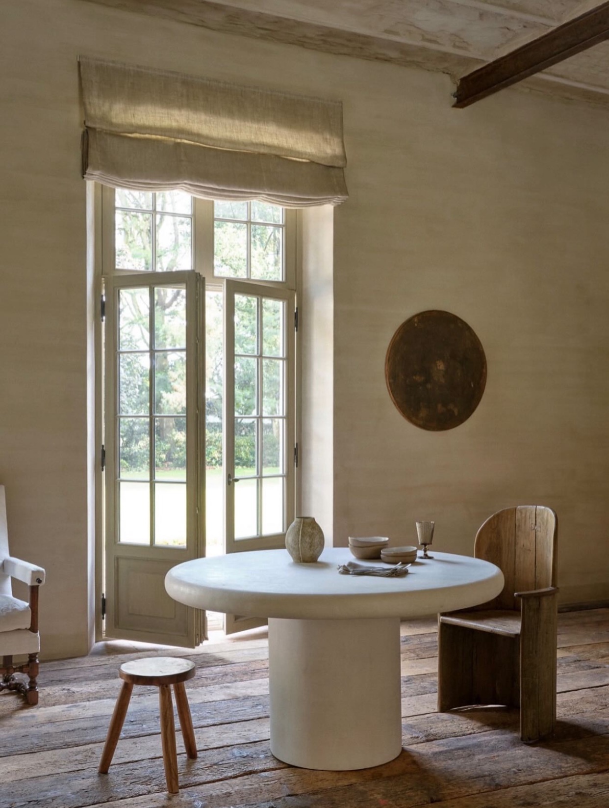 A minimalist interior with a round table, wooden chair, and large window. The room is bathed in natural light, creating a serene and inviting atmosphere.