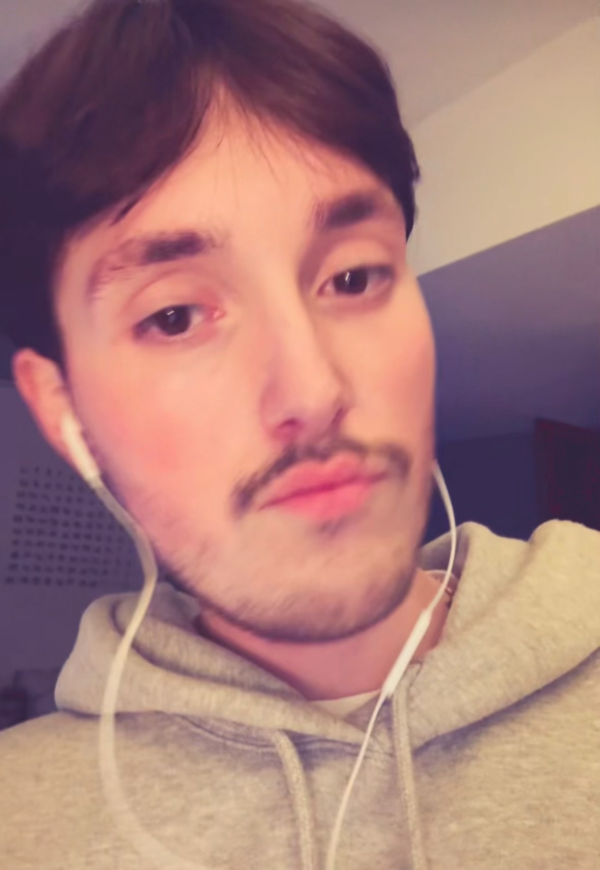 A close-up selfie of a young man with dark hair, stubble, and a mustache. He is wearing earbuds and a grey hoodie.