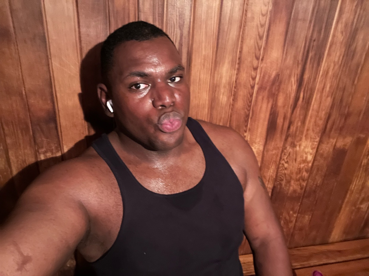 A man in a black tank top puckers his lips while sitting in a wooden sauna. He is wearing white earbuds and appears to be sweating.