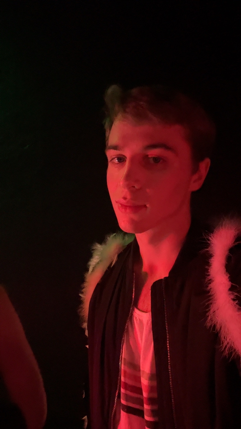 A young man poses for a selfie in a dimly lit room with red lighting. He is wearing a jacket and a white tank top.