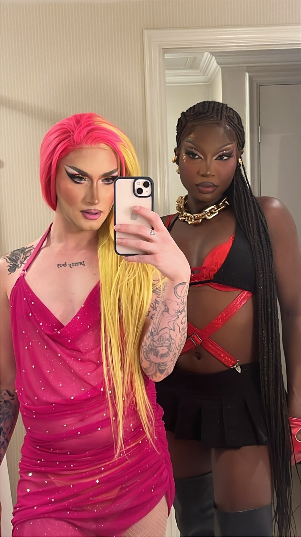 Two drag queens pose for a selfie, one with vibrant pink and yellow hair and a sparkling pink dress, the other with long braids, a black skirt, and a red and black top.