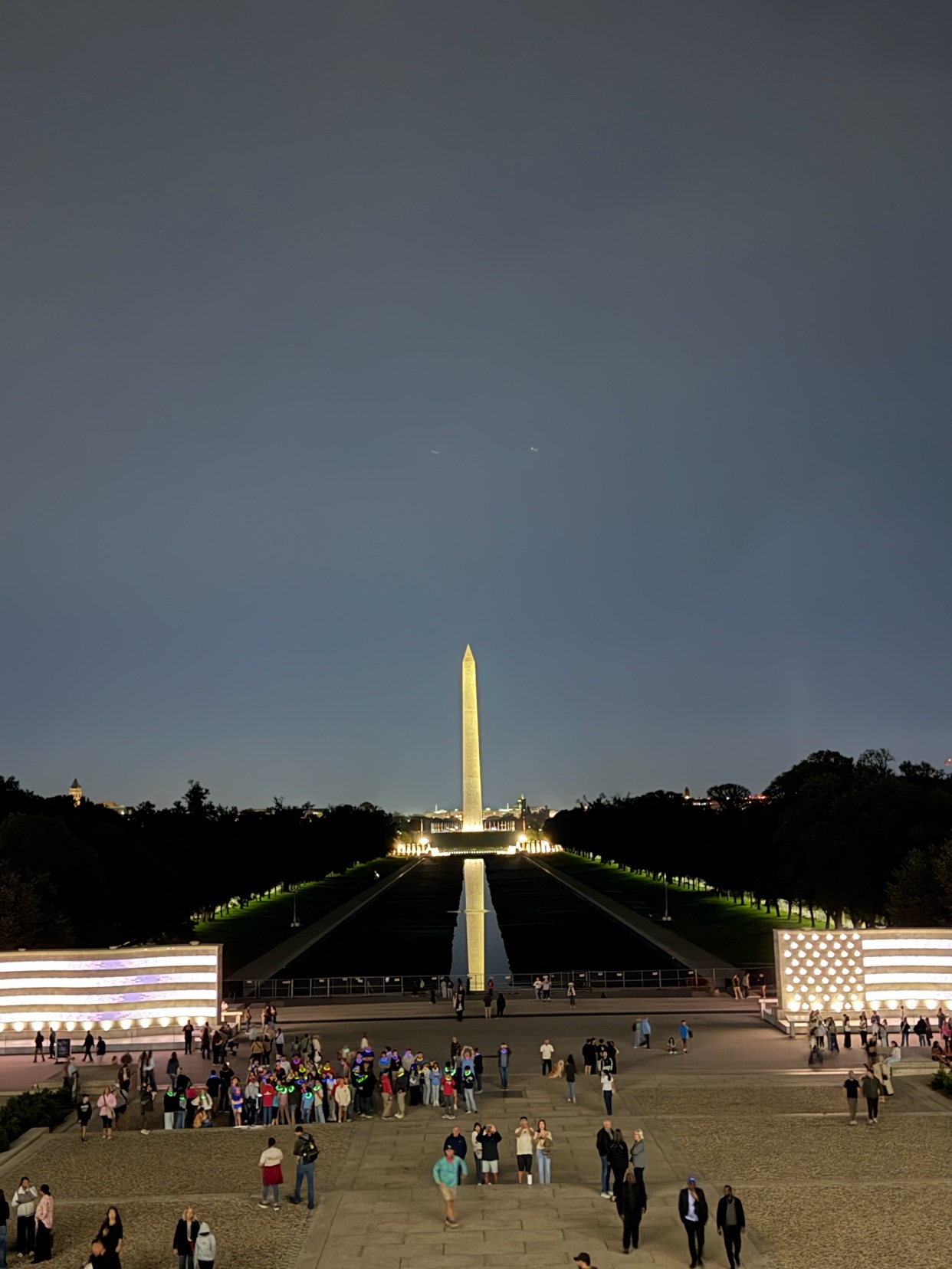 The Washington Monument stands tall against a twilight sky, illuminated and reflected in the Reflecting Pool. Crowds gather on the National Mall, with large flag displays flanking the scene.