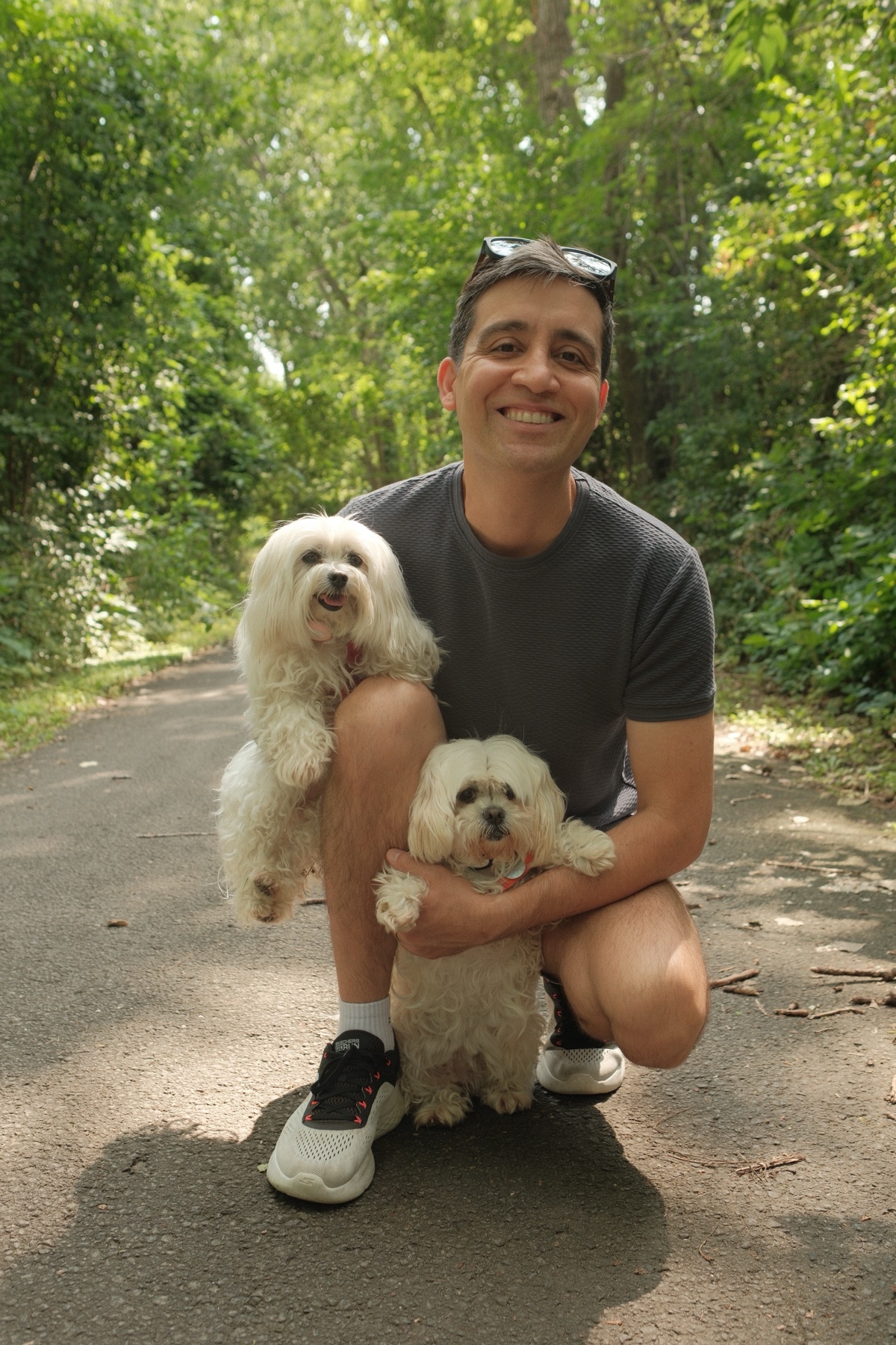 A man smiles while posing with two small white dogs on a path in a park. The man is wearing sunglasses on his head and sneakers.