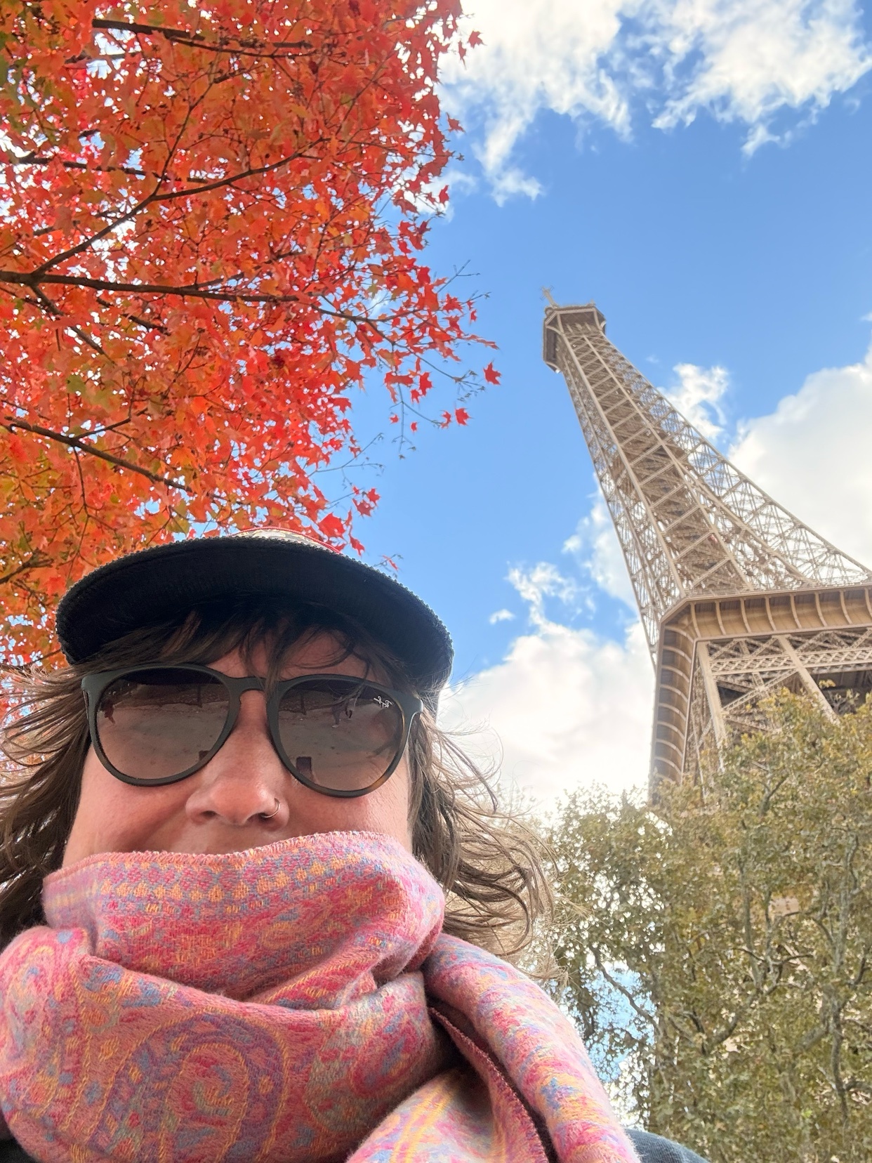 A woman takes a selfie in front of the Eiffel Tower in Paris, with vibrant red fall foliage framing the shot.