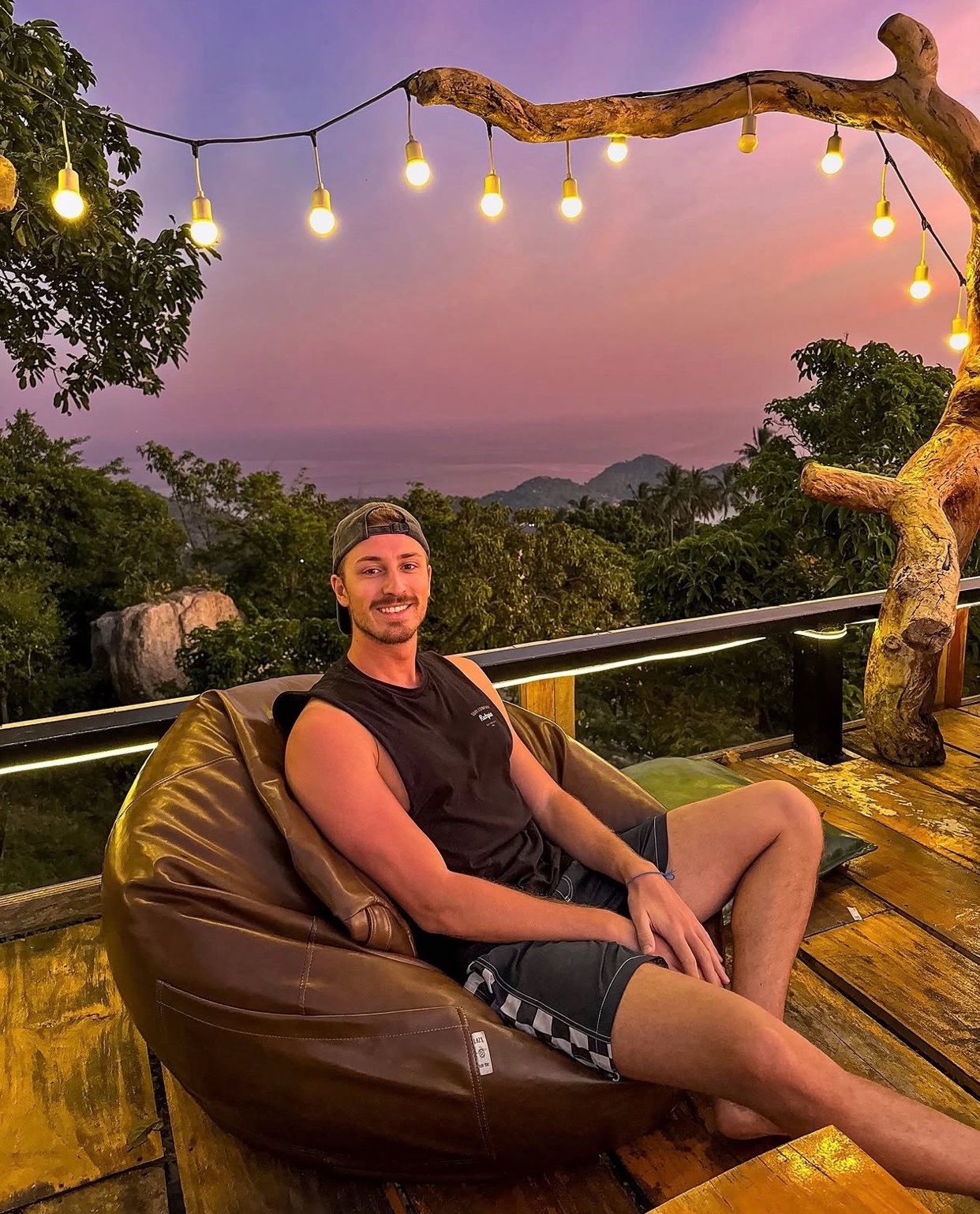 A man with stubble and a moustache sits in a large brown bean bag chair on a wooden deck. He wears a black tank top and checkered shorts, with a baseball cap on his head. String lights hang from a tree branch above, illuminating the tropical landscap
