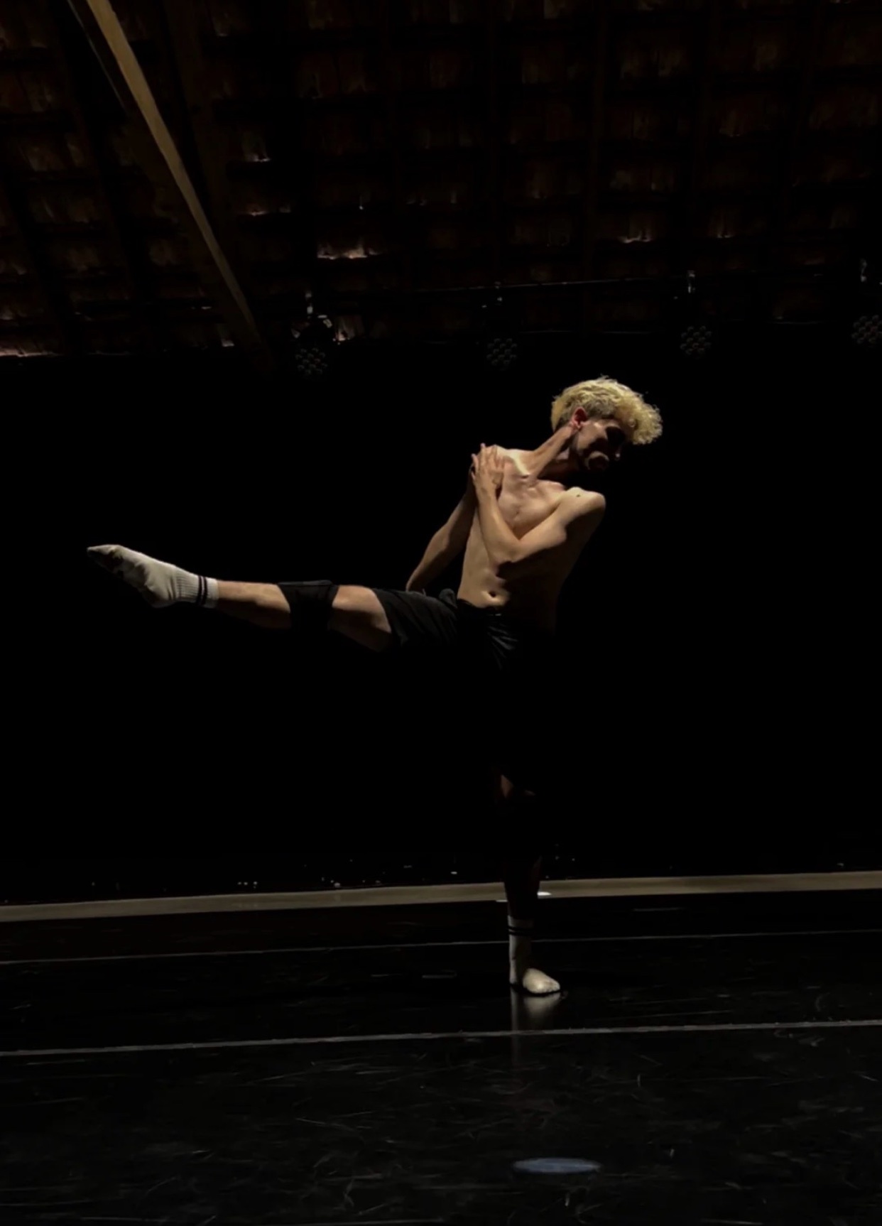 A shirtless man with blond hair performs a graceful ballet pose against a dark background. He is captured mid-motion, showcasing his athletic physique and artistic expression.
