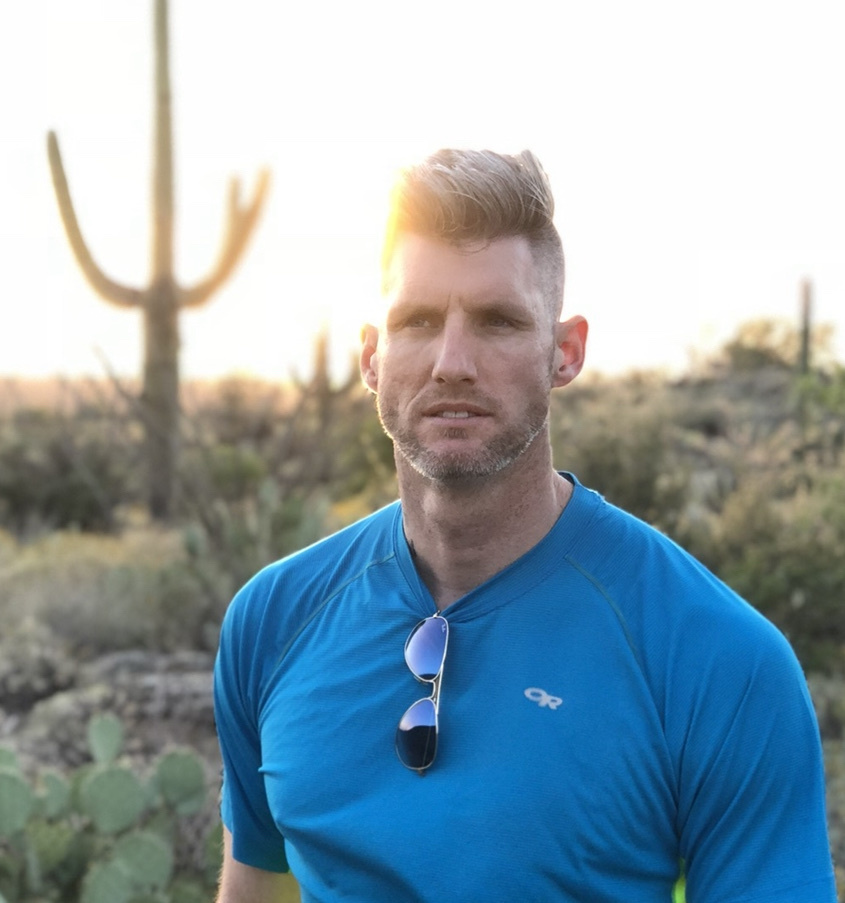 A man with short, styled hair and stubble poses in a desert landscape at sunset. He wears a bright blue athletic shirt with sunglasses hanging from the collar.