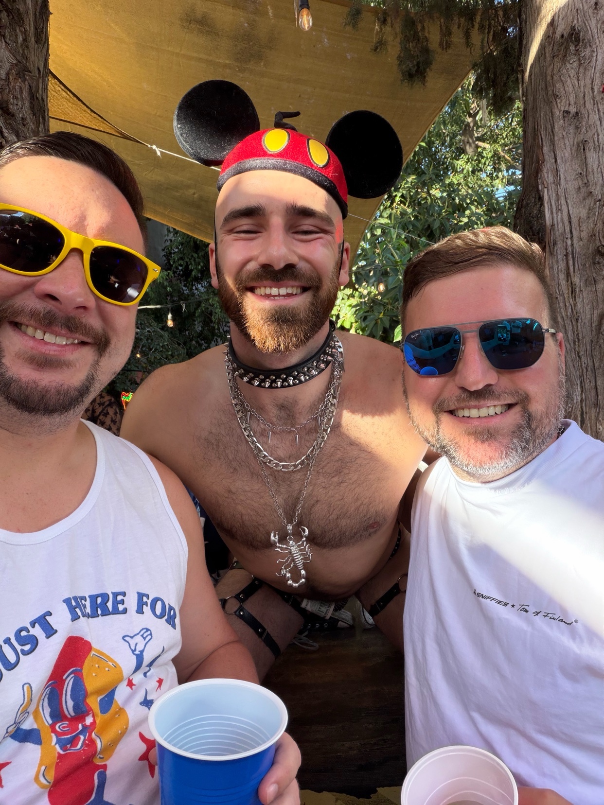 Three men smile for a selfie at an outdoor party. The man in the center wears Mickey Mouse ears and a leather harness, while the other two men wear t-shirts and sunglasses.
