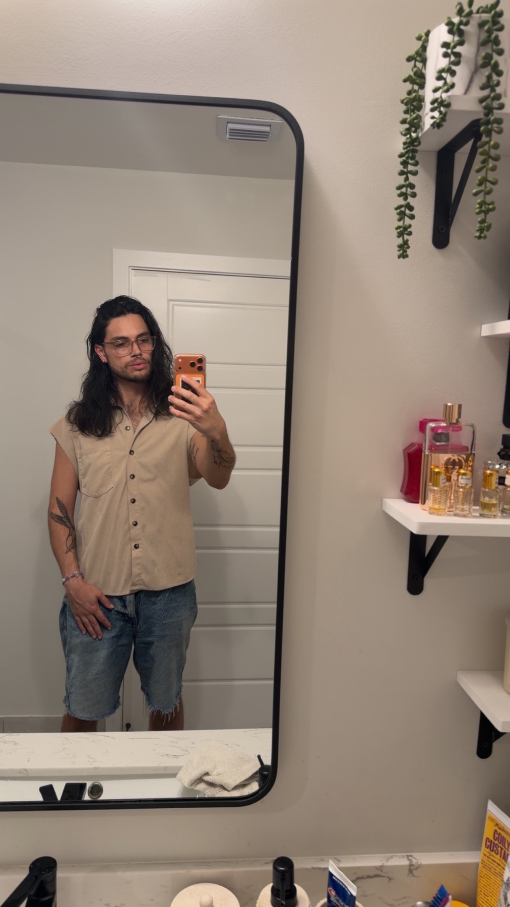 A man with long, dark hair and glasses takes a mirror selfie in a bathroom. He is wearing a tan button-up shirt and denim shorts.