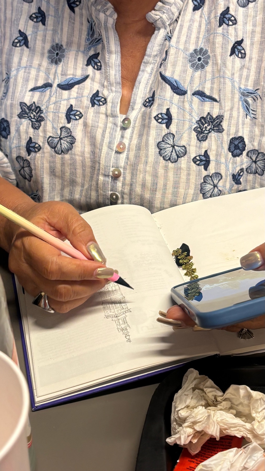 A woman with a floral and striped shirt is writing in a notebook while holding a phone. Her nails are painted and she is wearing a ring.