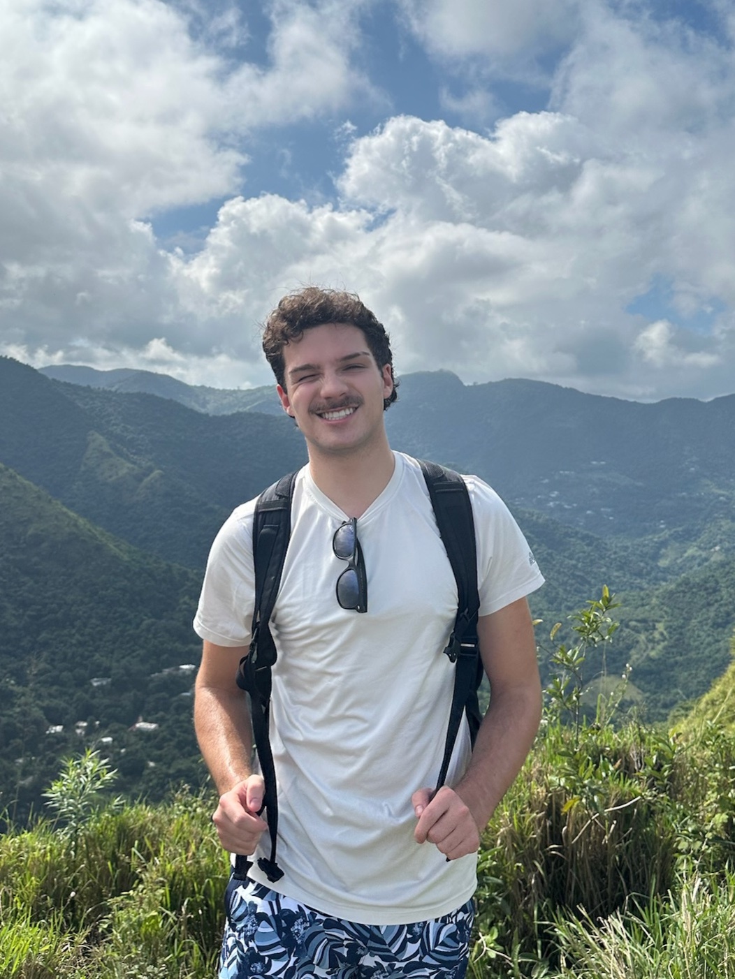 A man smiles at the camera while standing on a mountain. He is wearing a white t-shirt and a backpack.