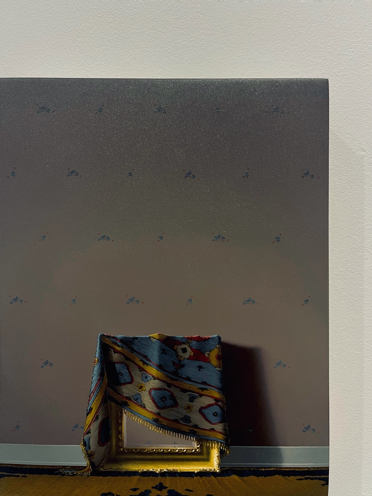 A painting depicts a still life of a framed artwork draped with a colorful tapestry against a patterned wallpaper background.