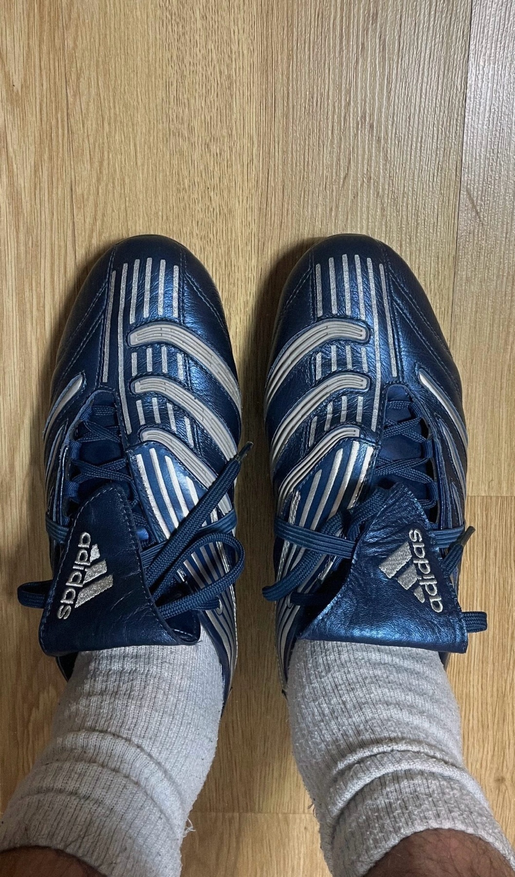 A top-down shot of a pair of blue and white Adidas soccer cleats. The shoes are placed on a wooden floor.