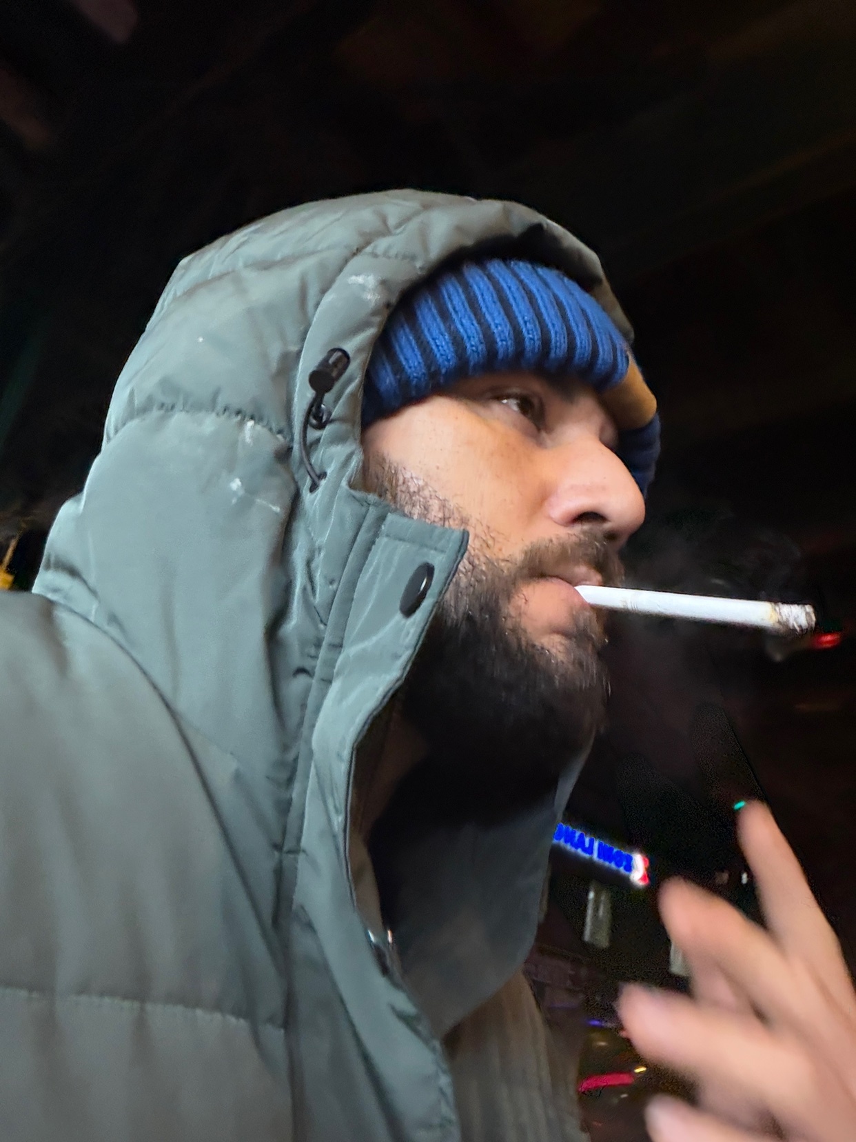 A man is smoking a cigarette while wearing a winter coat and beanie. The photo is taken at night.