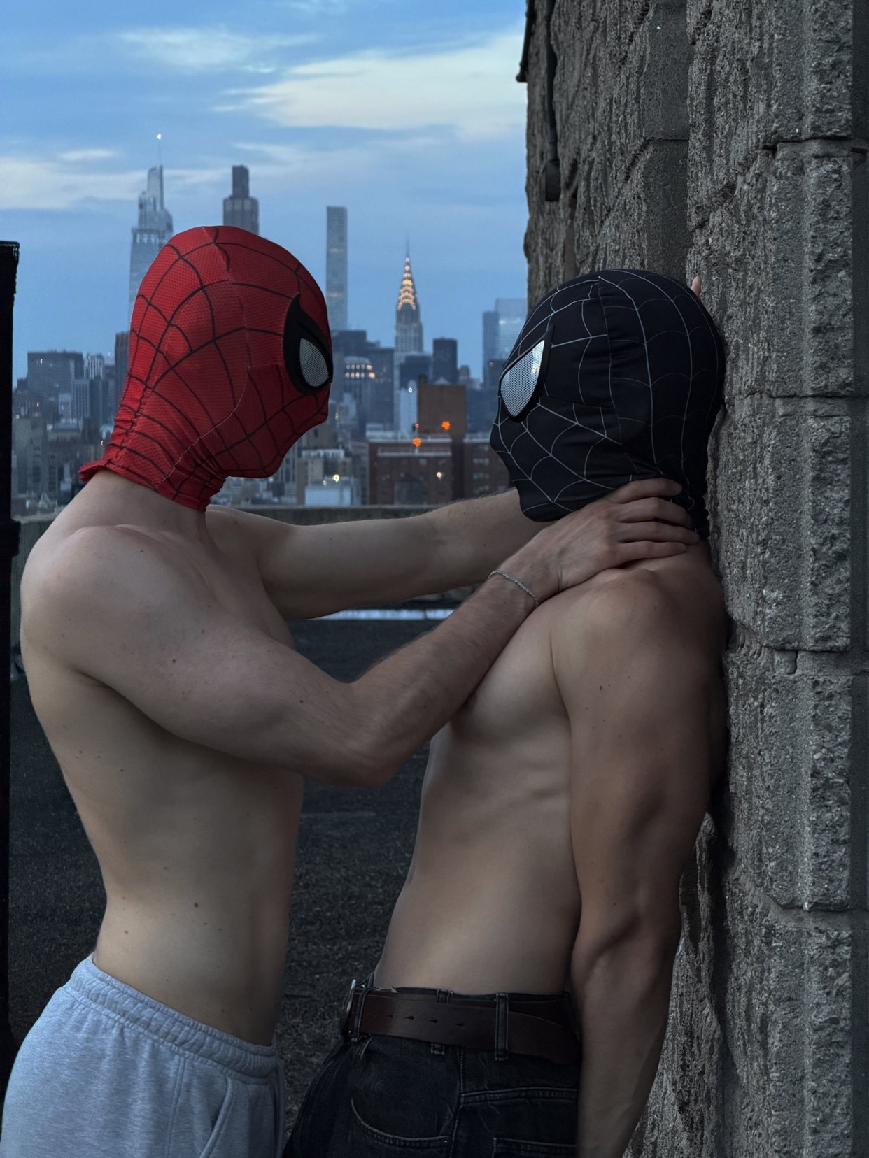 Two men in Spiderman masks lean against a brick wall, with the New York City skyline in the background. Both men are shirtless, with one gently touching the other's neck.