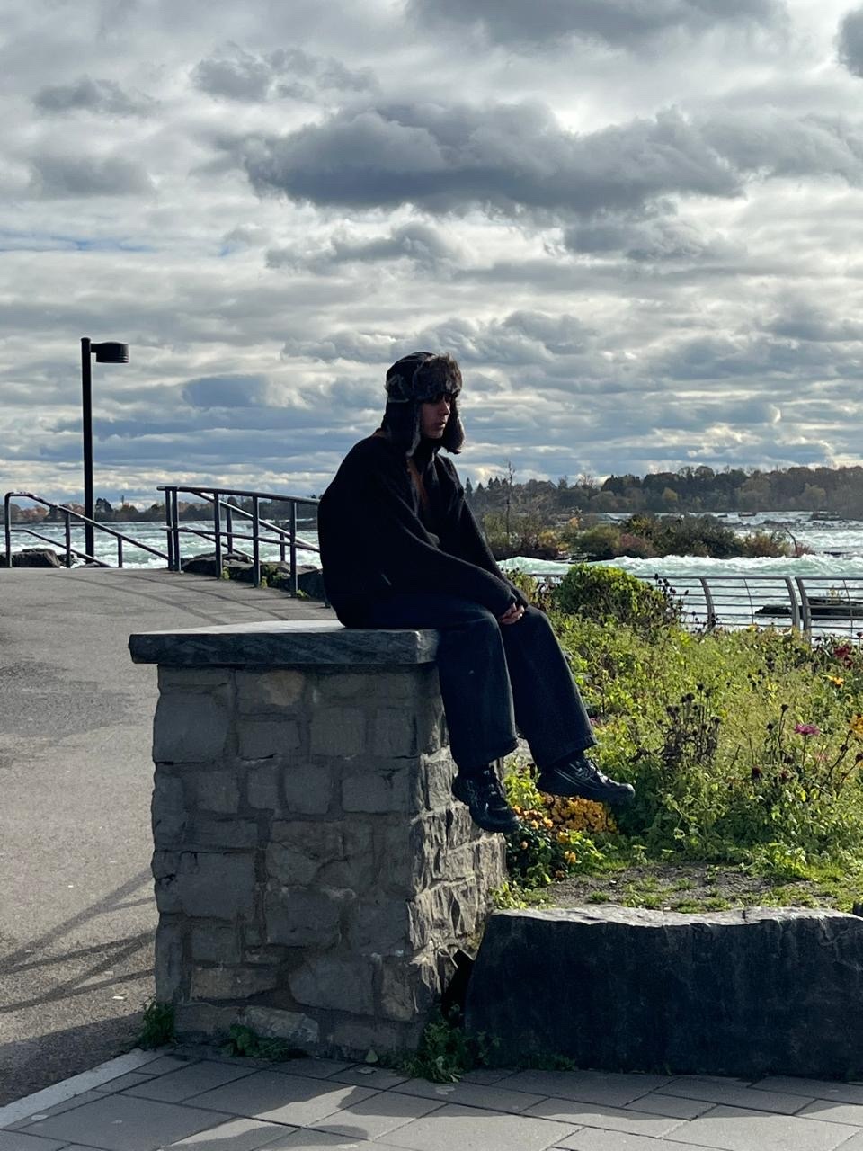 A woman sits on a stone wall, overlooking Niagara Falls. She is wearing a black outfit and a hat.