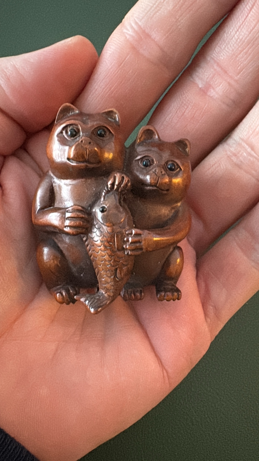 A close-up shot of a person's hand holding a small, intricately carved Japanese netsuke depicting two cats holding a fish. The netsuke appears to be made of wood or a similar material and has a warm, brown patina.
