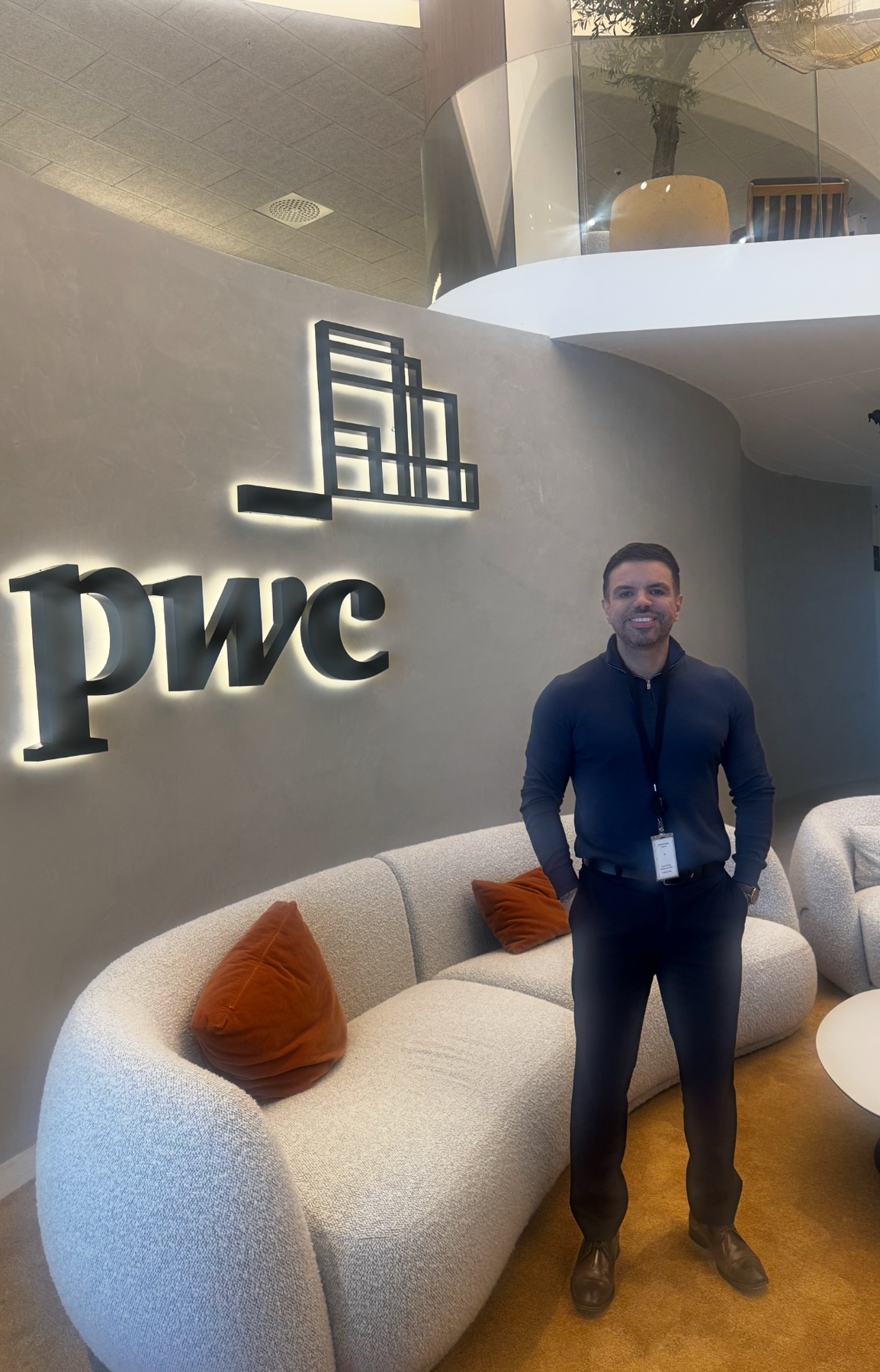 A man stands in front of the PwC logo in a modern office setting. He is smiling and wearing a suit.