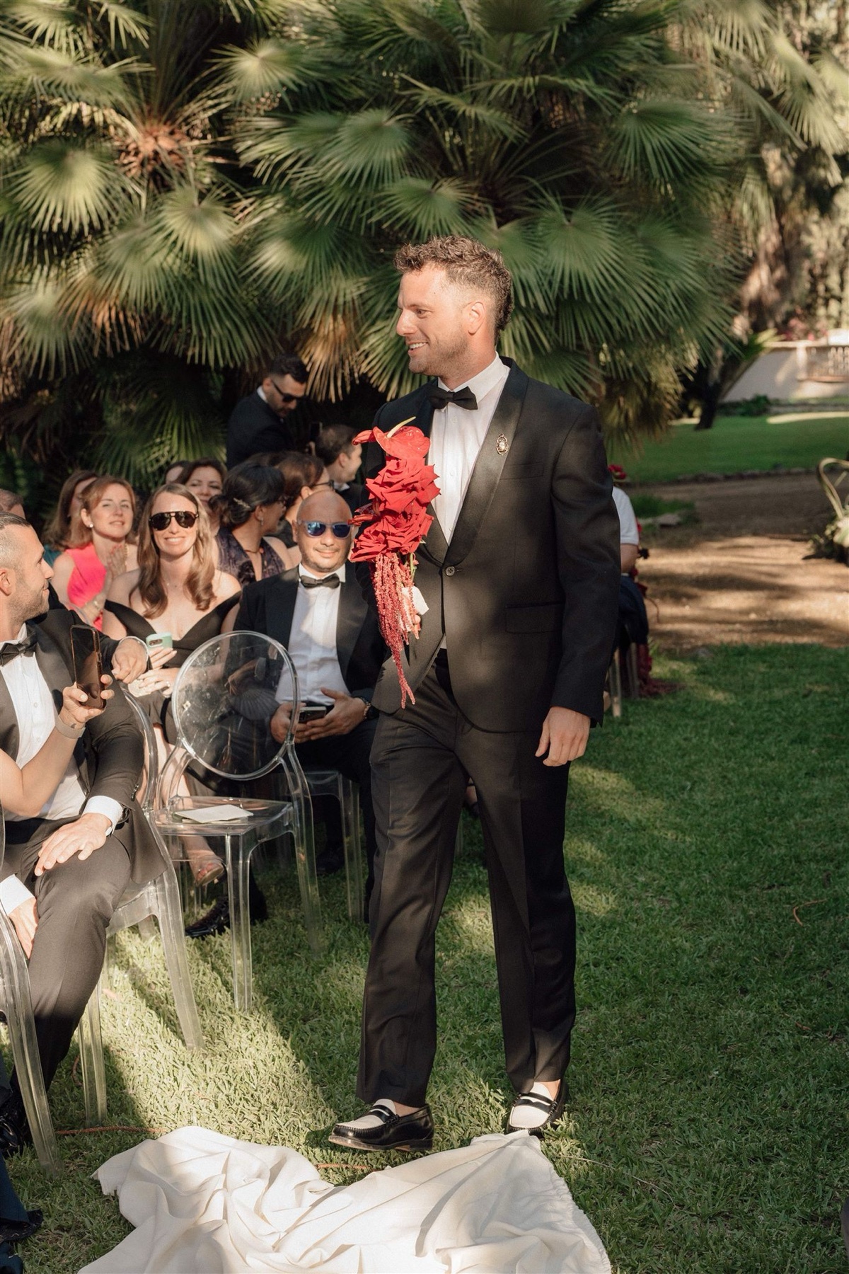 A man in a black suit walks down an aisle at an outdoor wedding. He is holding a red bouquet and smiling.