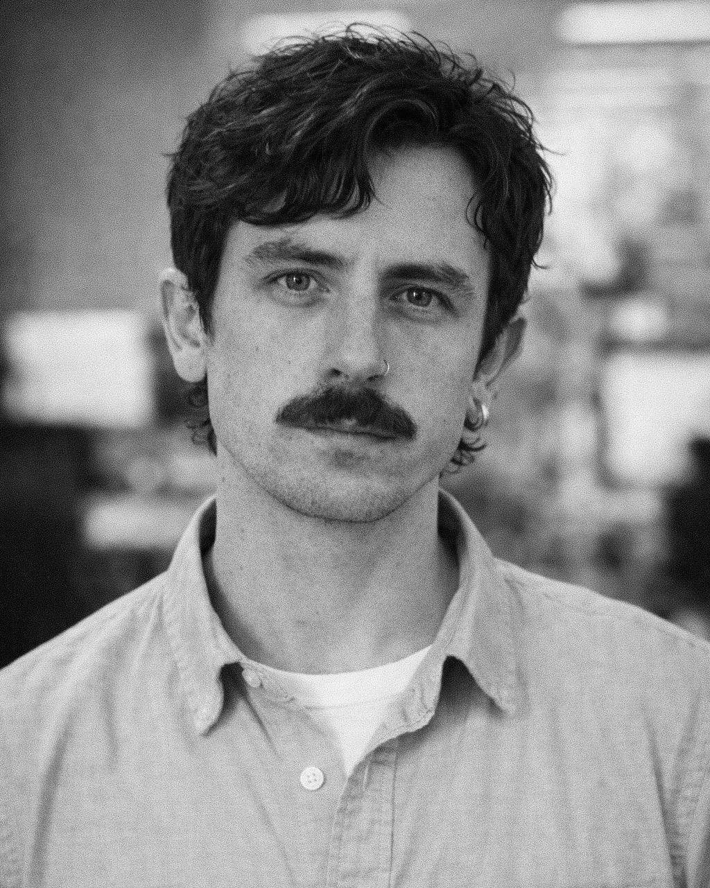A black and white headshot of a man with dark hair and a mustache. He is wearing a button-down shirt and looking directly at the camera.