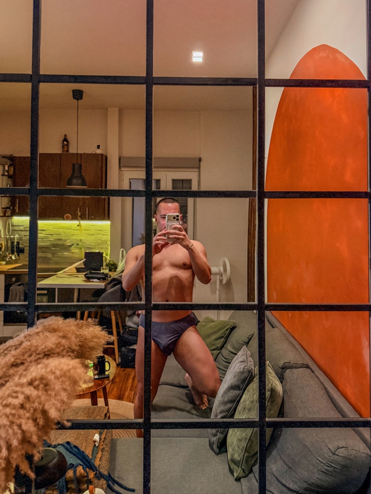 A man takes a mirror selfie, shirtless and in underwear. The photo is taken in a modern apartment with an orange wall decor.