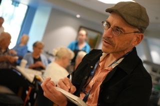 A man wearing glasses and a flat cap speaks to a group of people in the background. He is holding a book or pamphlet and appears to be in the midst of a discussion.