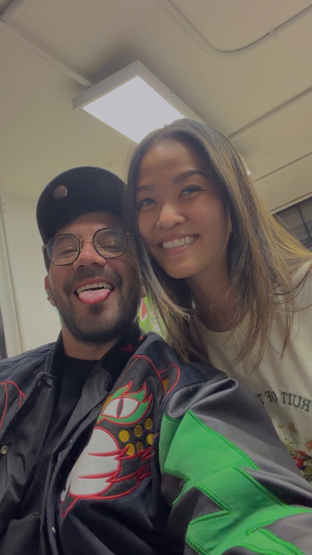 A man and a woman smile for a selfie. The man, wearing glasses and a baseball cap, sticks his tongue out playfully. The woman has long, brown hair and is wearing a light-colored t-shirt.
