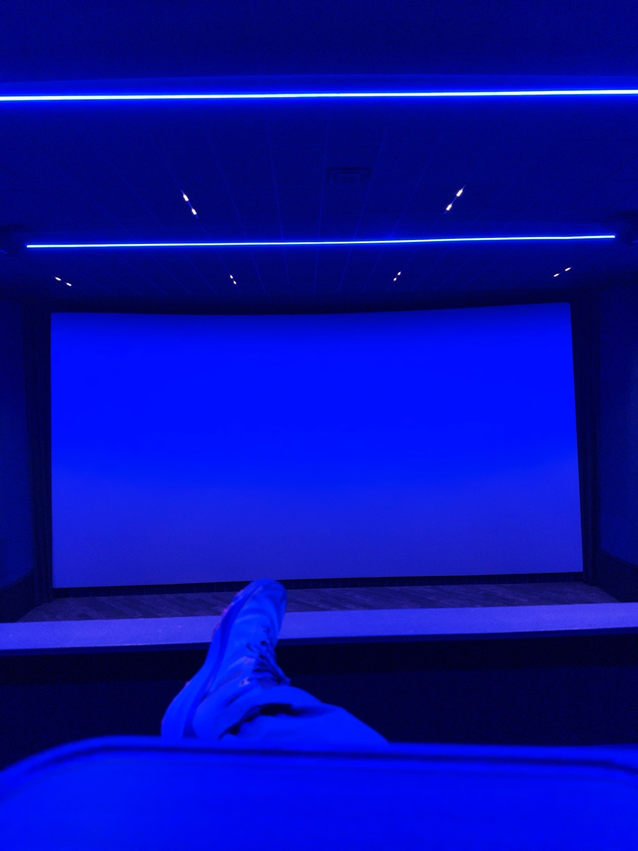 A person's legs and feet in white sneakers are visible in the foreground, pointing towards a large, blank movie screen in a dark theater. The room is illuminated by vibrant blue LED lighting along the ceiling and walls, creating a futuristic ambiance