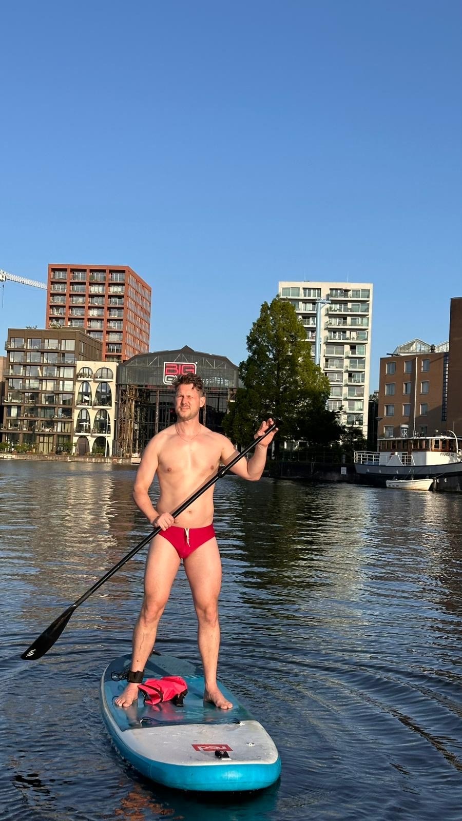 A shirtless man in red swimwear stands on a paddleboard, holding a paddle. He is on a body of water with buildings and trees in the background under a clear blue sky.