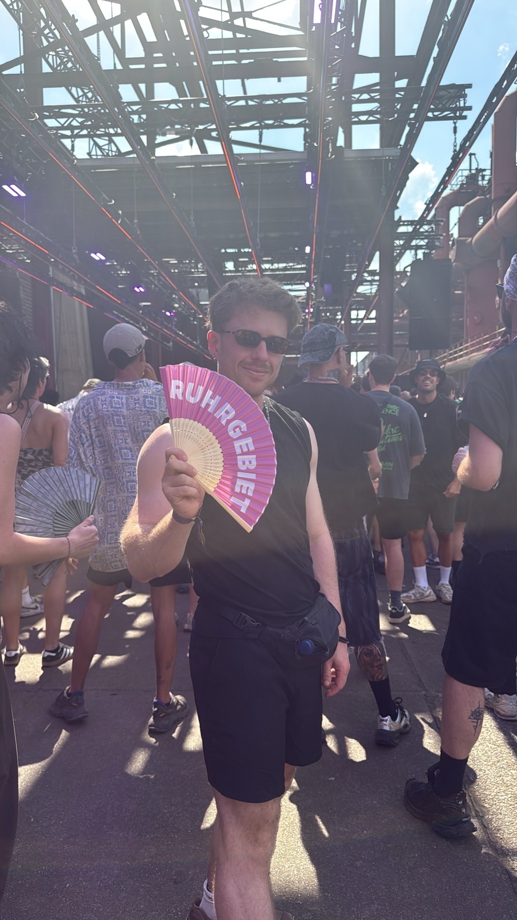 A man in sunglasses holds a pink fan that says "RUHRGEBIET" in front of a crowd at an outdoor event. The background features industrial structures and a bright, sunny sky.