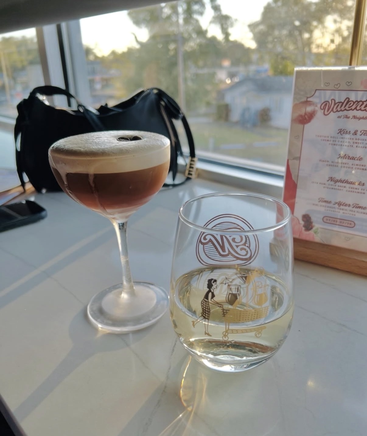 A close-up shot of two drinks, an espresso martini and a glass of white wine, sit on a table near a window. A drink menu with a Valentine's Day theme is visible in the background.