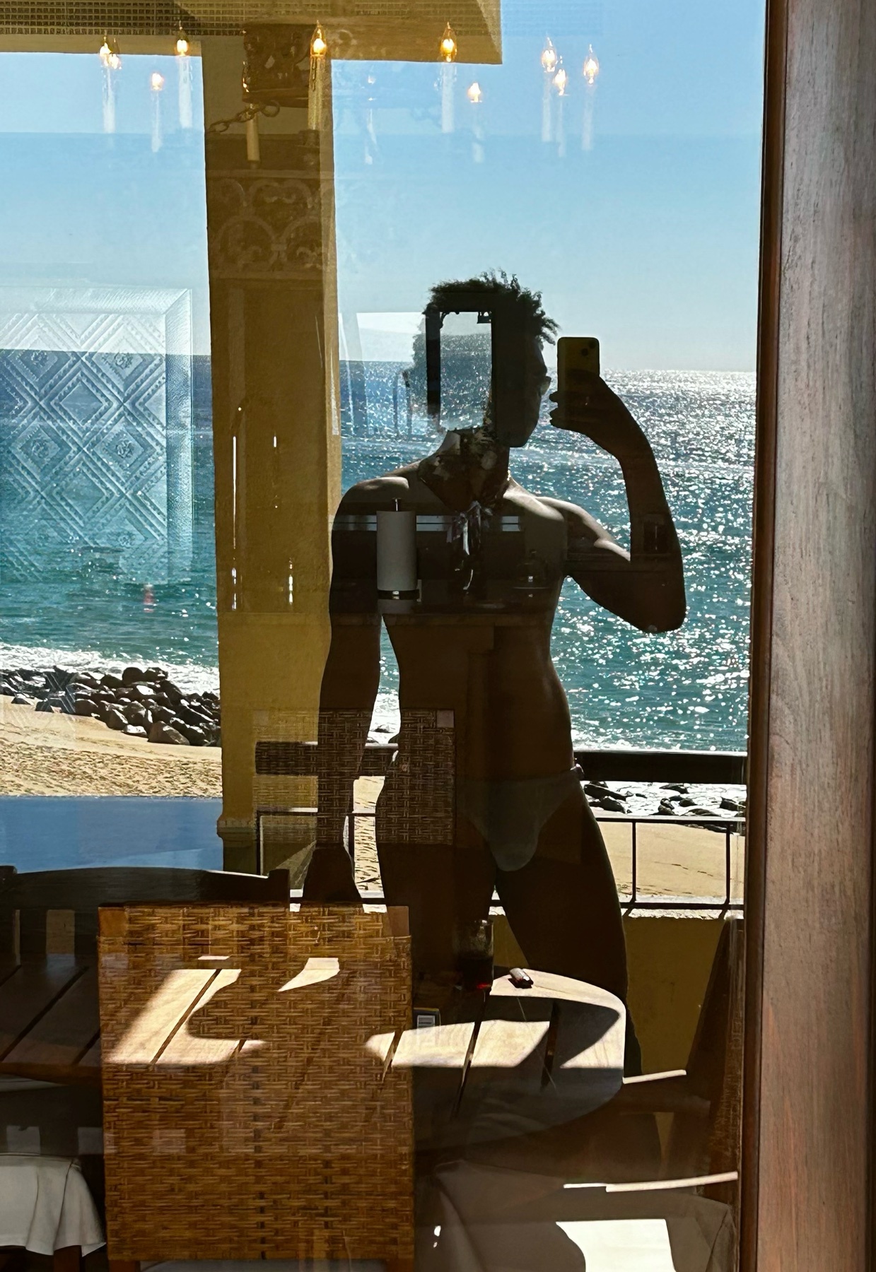 A man takes a shirtless selfie in front of a window, with a beach and ocean view in the background. The reflection shows his body and the sunny outdoor scene.