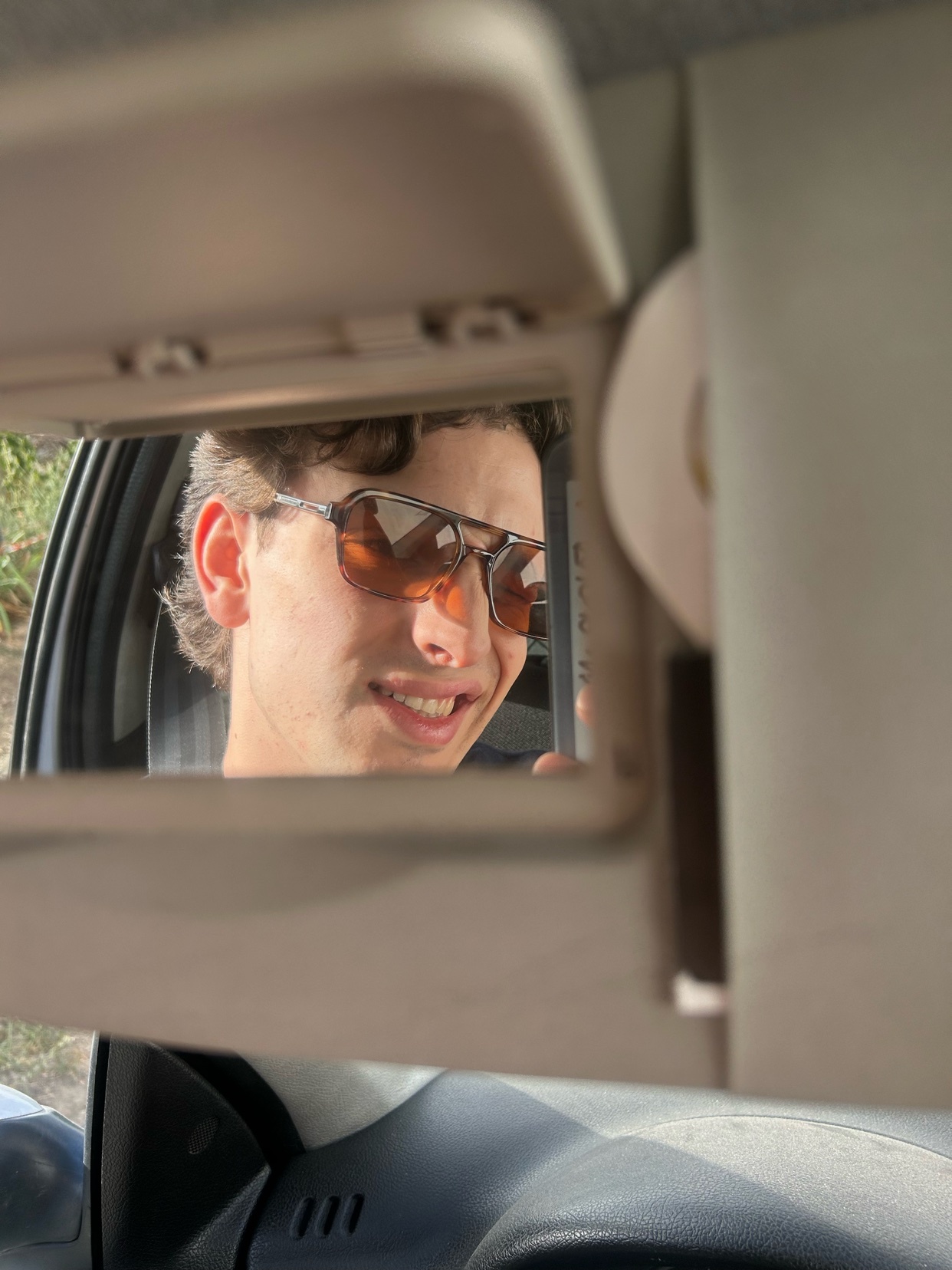 A man takes a selfie in a car mirror, wearing sunglasses and smiling. The image is a close-up, focusing on the man's face and the reflection.