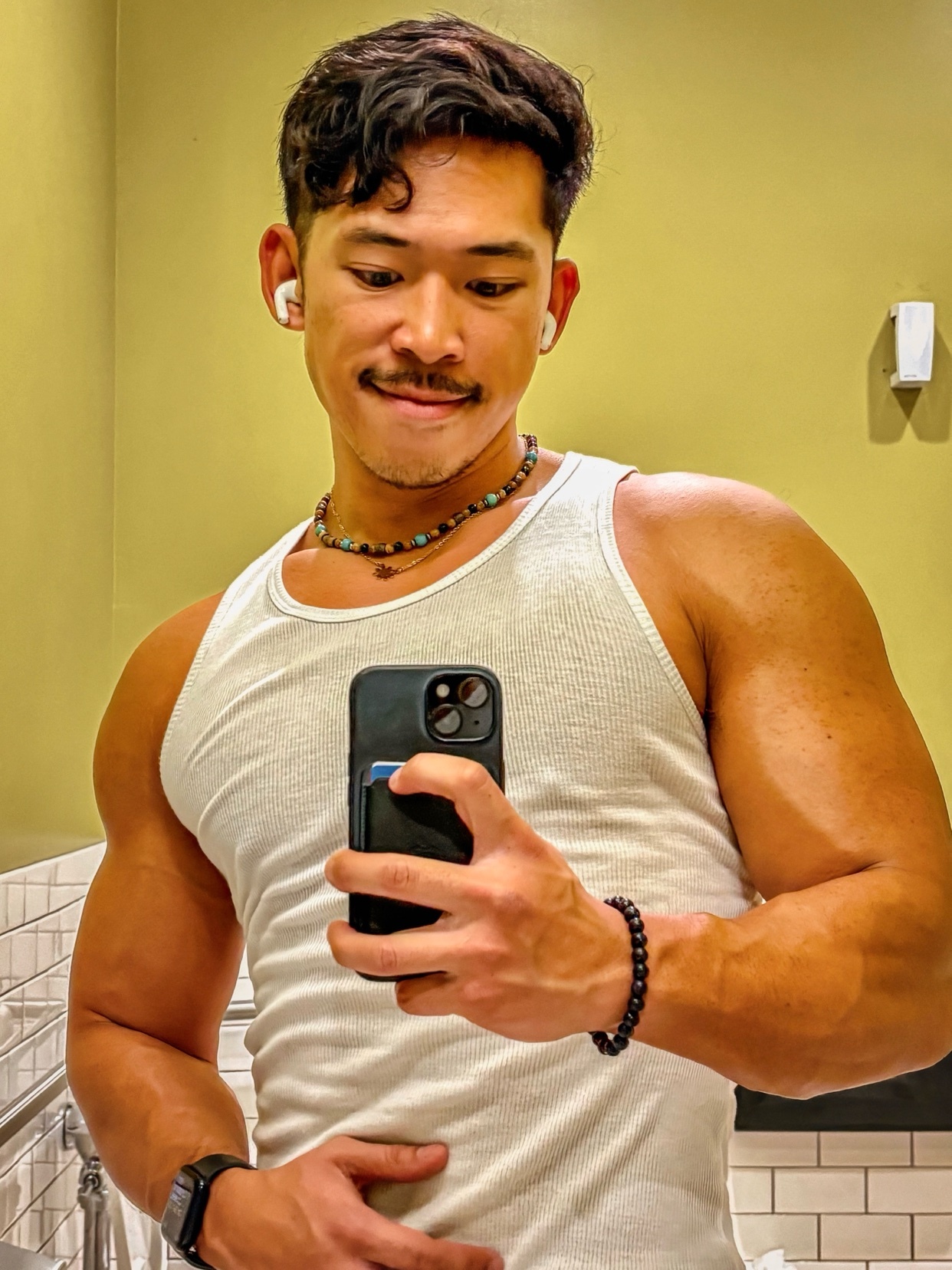 A man takes a mirror selfie, showcasing his physique. He is shirtless, flexing his arm muscles.
