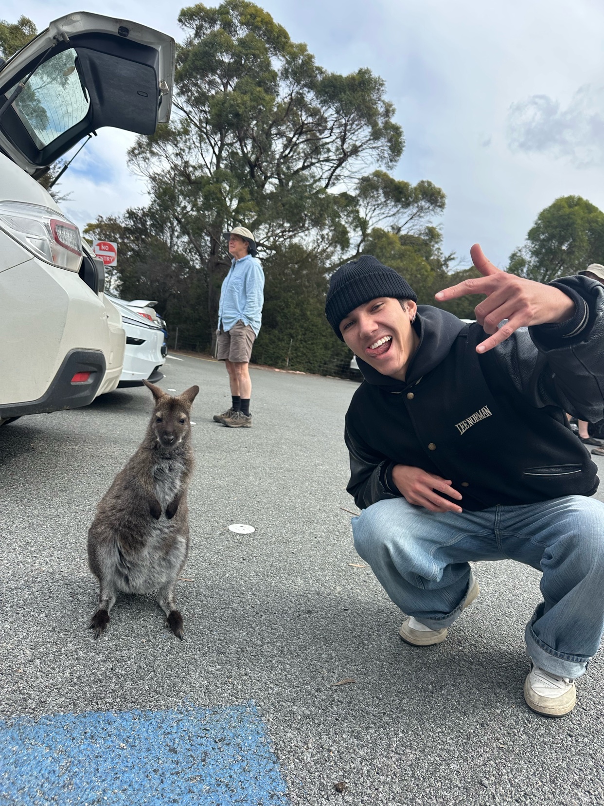 A man in a black beanie and jeans crouches down next to a wallaby in a parking lot. Another man stands in the background near a car.