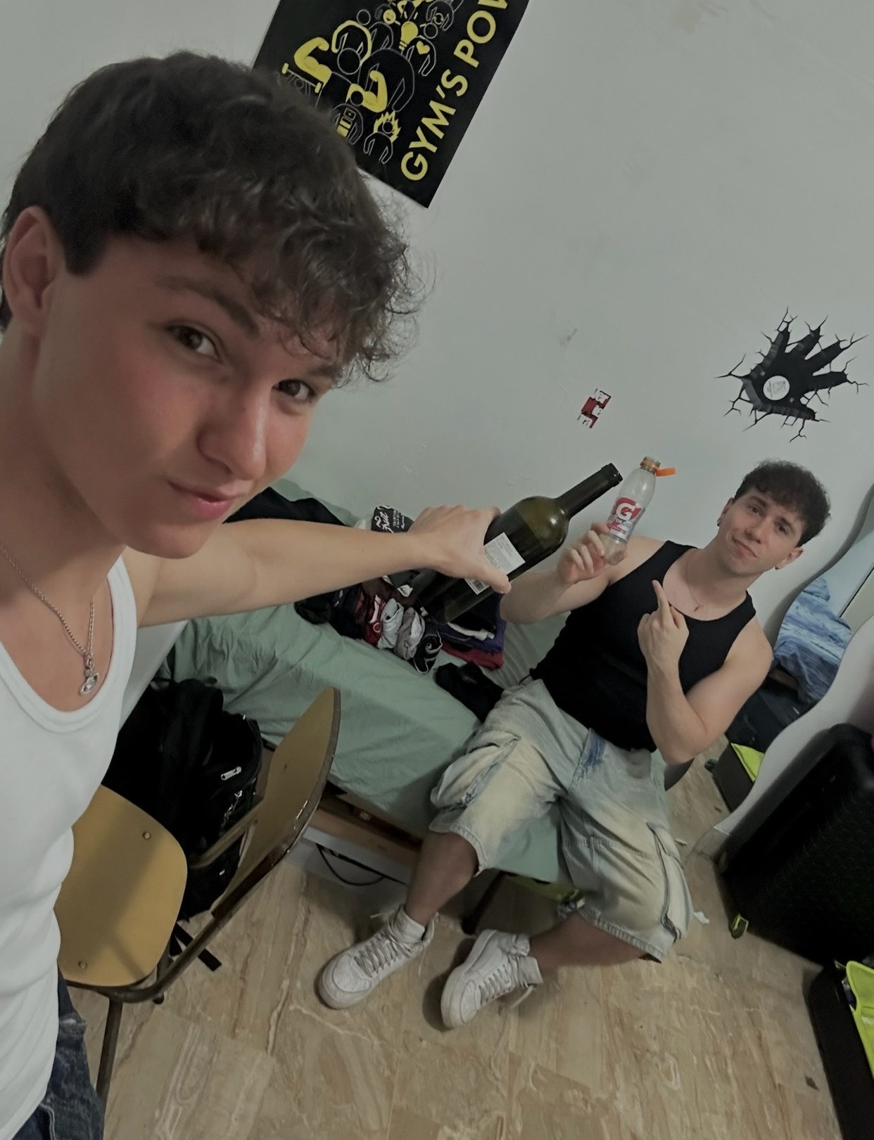 Two young men pose for a selfie, one holding a bottle of wine and the other a bottle of liquor. They are both shirtless and in a room.