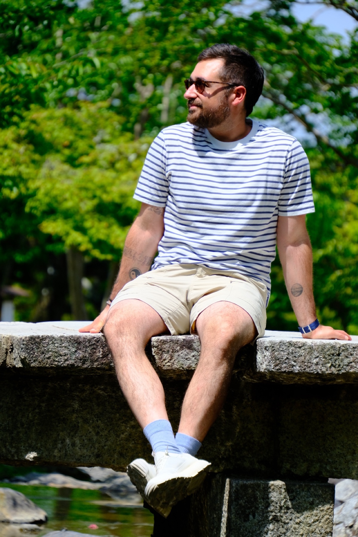 A man with a beard and sunglasses sits outdoors on a stone ledge, wearing a striped t-shirt and shorts. He is looking off to the side with lush green trees in the background.