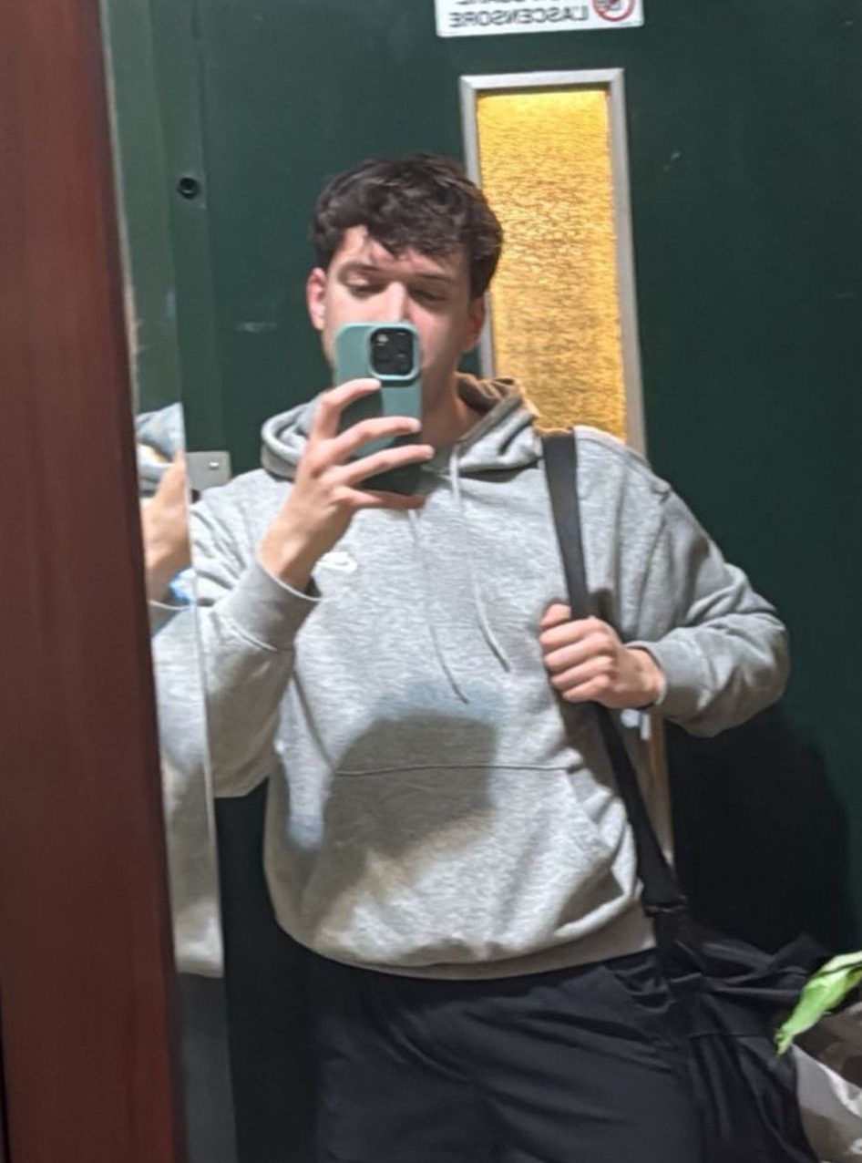 A man takes a selfie in a grey hoodie and black shorts, holding a gym bag over his shoulder.