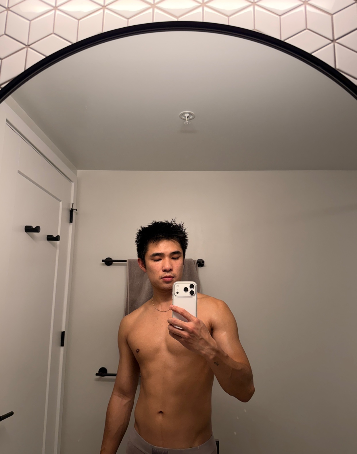 A man takes a shirtless selfie in a bathroom mirror. The photo highlights his physique and the modern design of the space.